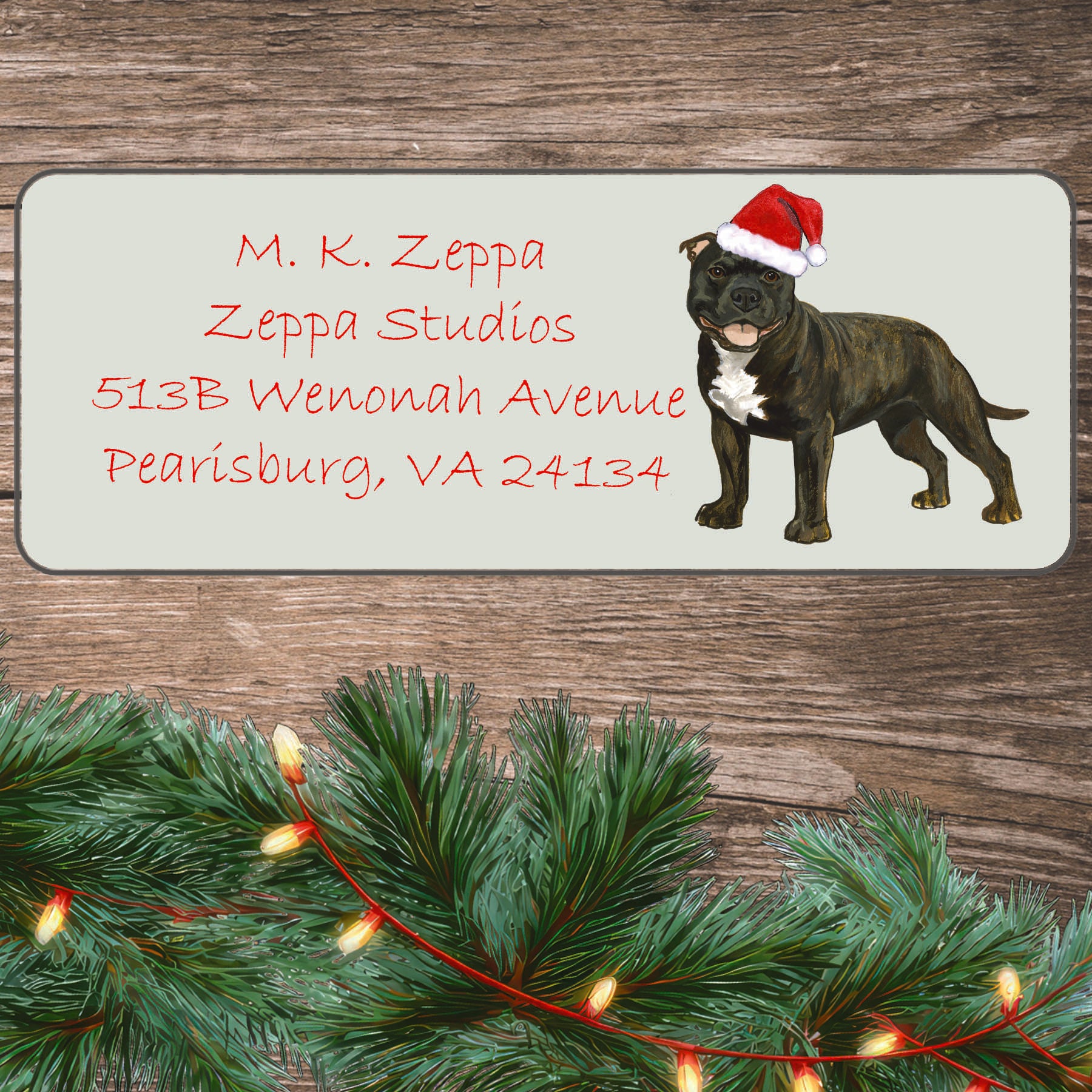 An illustration of a brindle English Staffordshire Terrier wearing a Santa Hat on an ivory return address label set off by a wood surface with holiday greenery background.