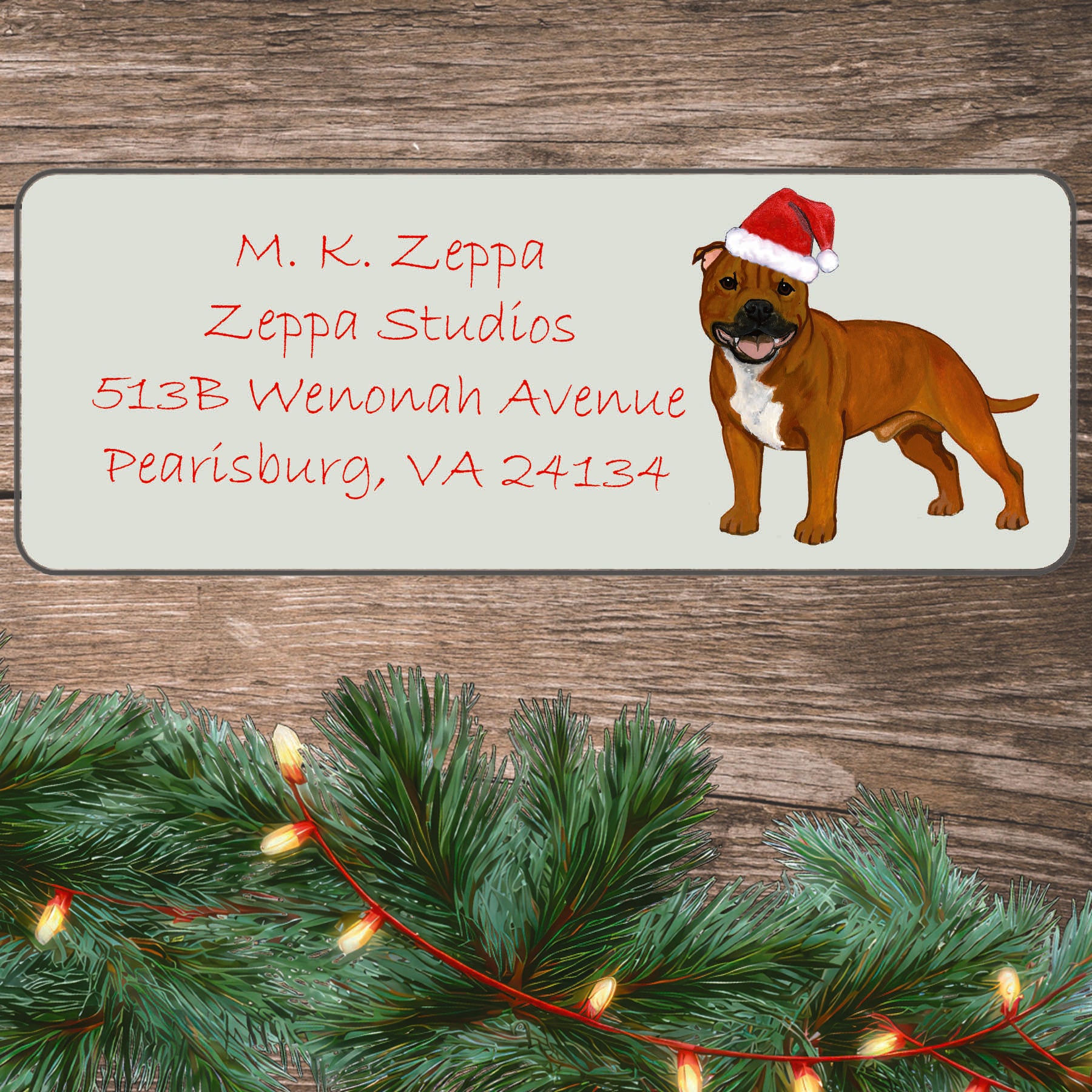 An illustration of a red English Staffordshire Terrier wearing a Santa Hat on an ivory return address label set off by a wood surface with holiday greenery background.
