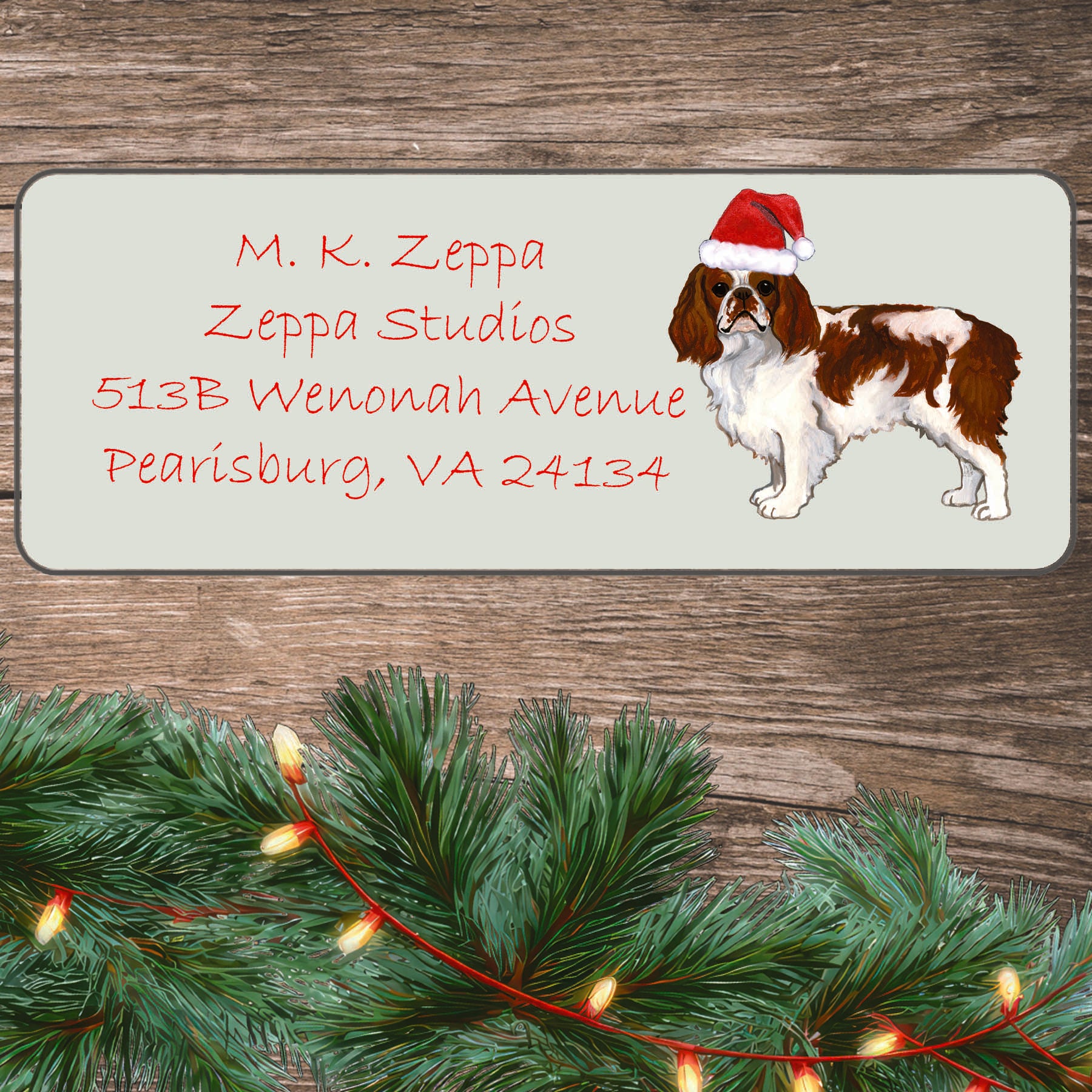 An illustration of a Blenheim English Toy Spaniel wearing a Santa Hat on an ivory return address label set off by a wood surface with holiday greenery background.
