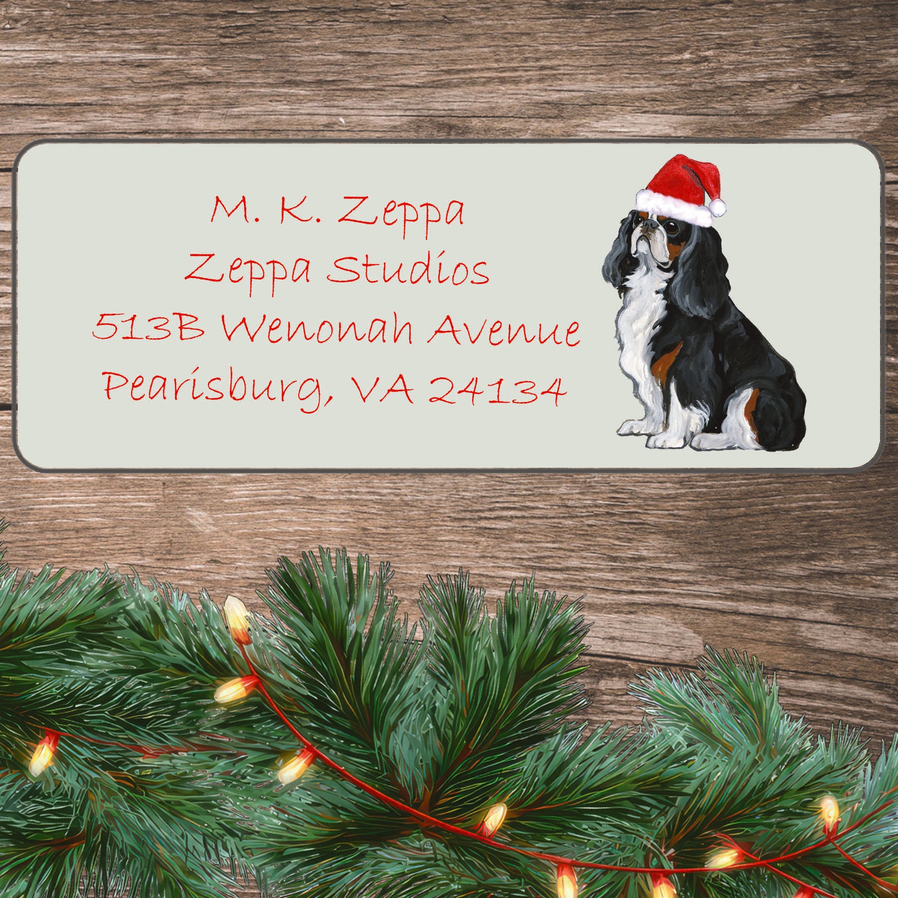 An illustration of a tri-color English Toy Spaniel wearing a Santa Hat on an ivory return address label set off by a wood surface with holiday greenery background.