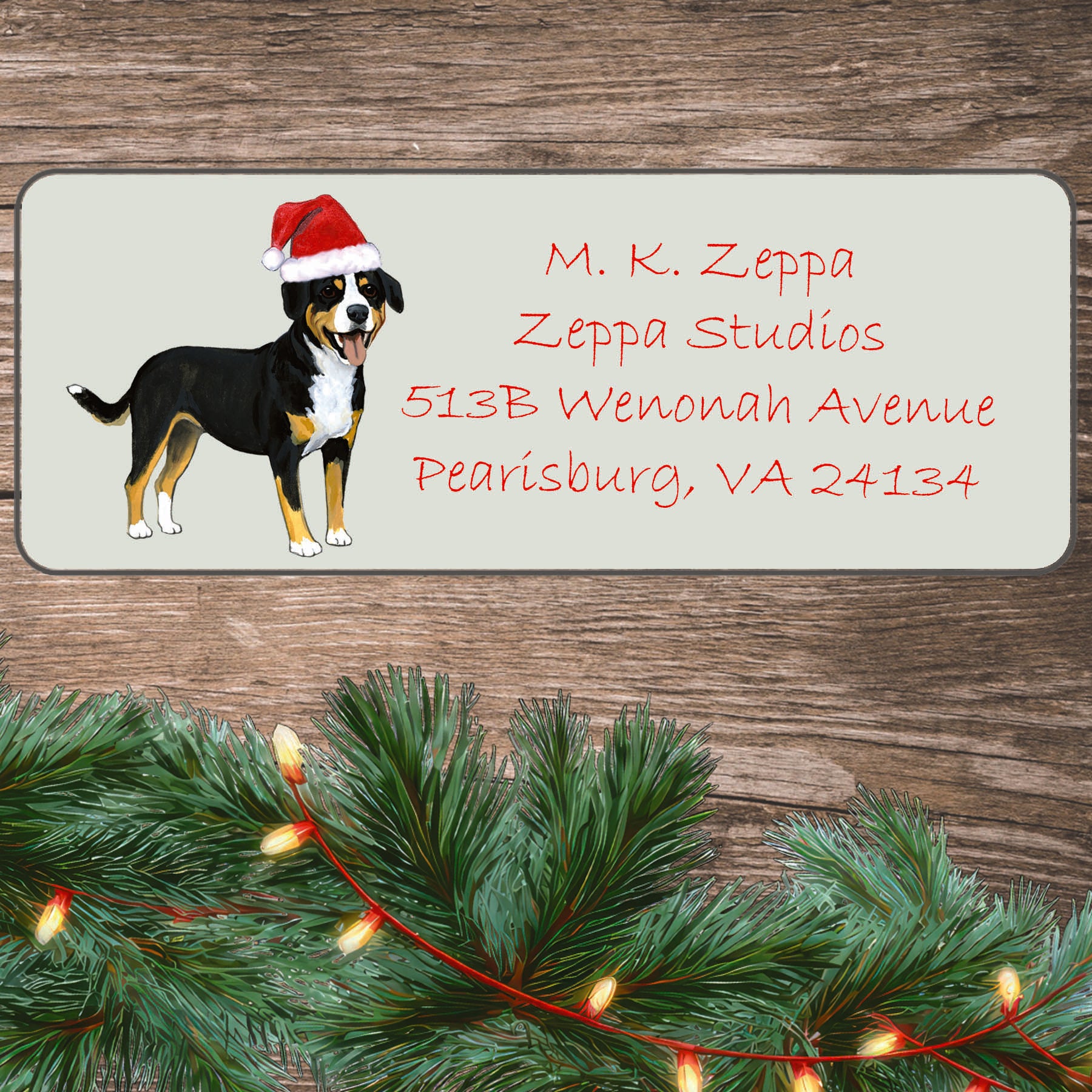 An Entelbucher Mountain Dog in a Santa Hat on an ivory return address label set off by a wood surface with holiday greenery background.