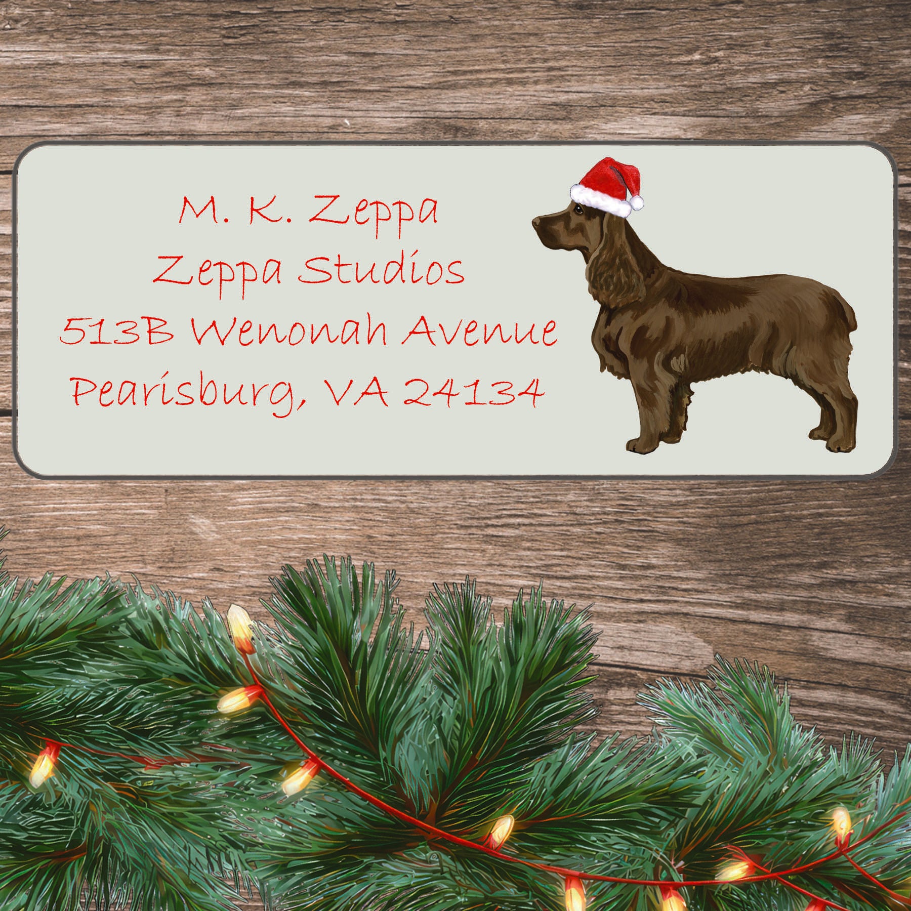 An illustration of a Field Spaniel wearing a Santa Hat on an ivory return address label set off by a wood surface with holiday greenery background.