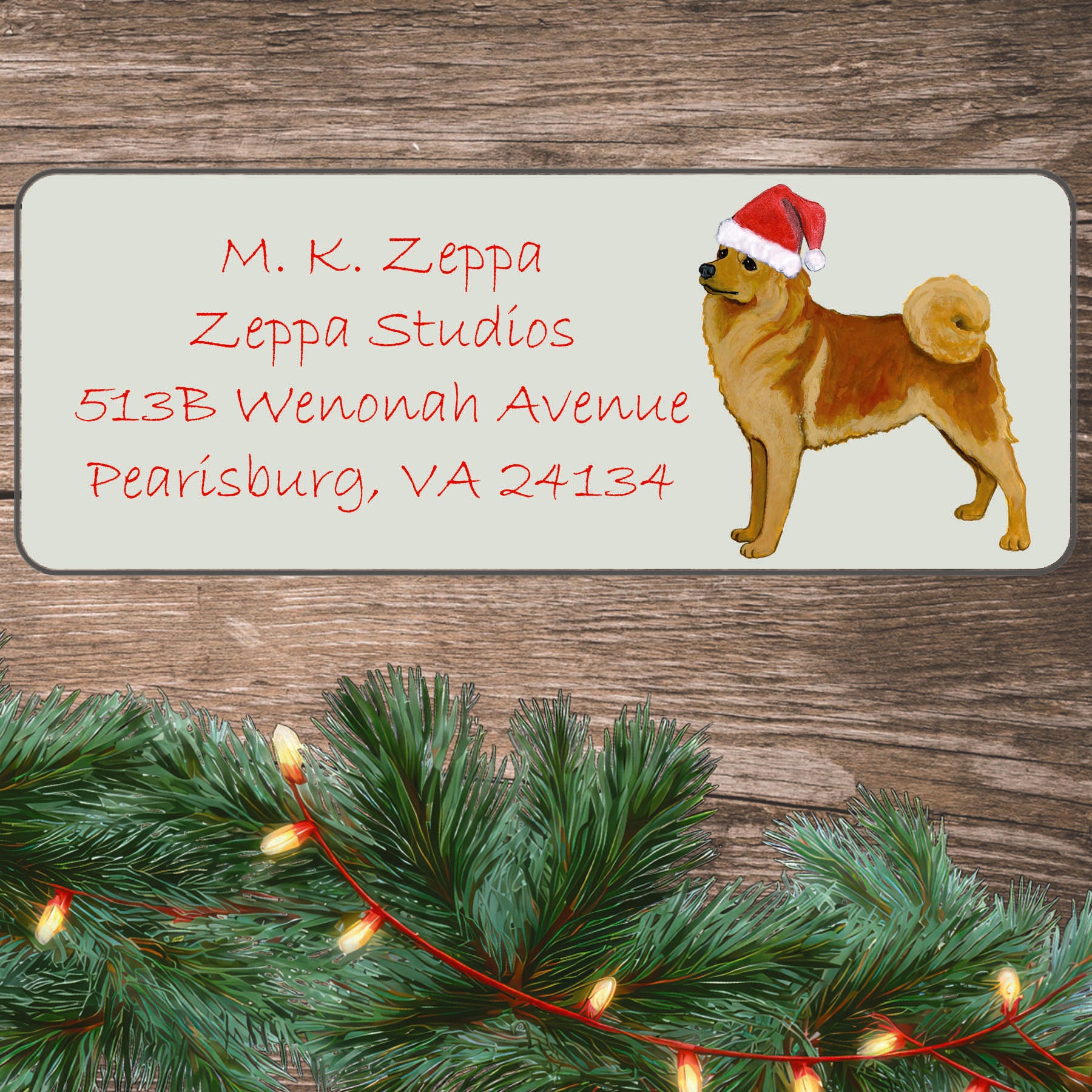An illustration of a Finnish Spitz wearing a Santa Hat on an ivory return address label set off by a wood surface with holiday greenery background.
