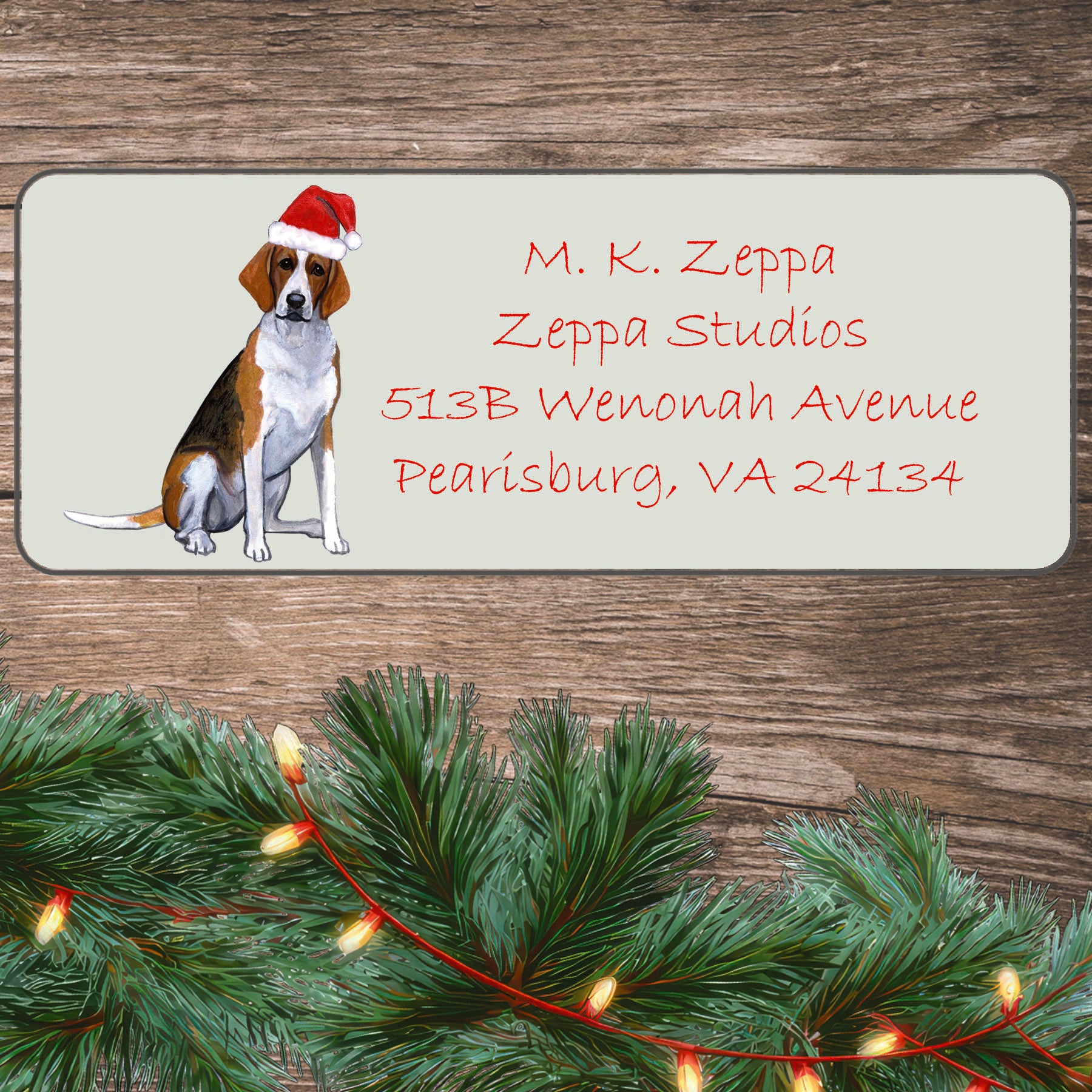 An illustration of a Foxhound wearing a Santa Hat on an ivory return address label set off by a wood surface with holiday greenery background.