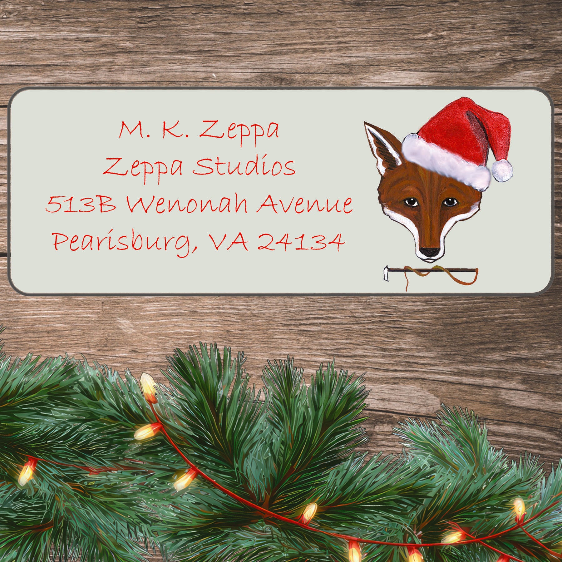 An illustration of a Fox face wearing a Santa Hat on an ivory return address label set off by a wood surface with holiday greenery background. 