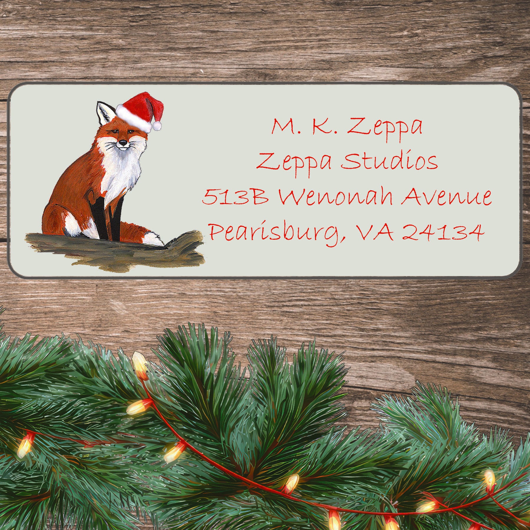 An illustration of a sitting Fox wearing a Santa Hat on an ivory return address label set off by a wood surface with holiday greenery background.