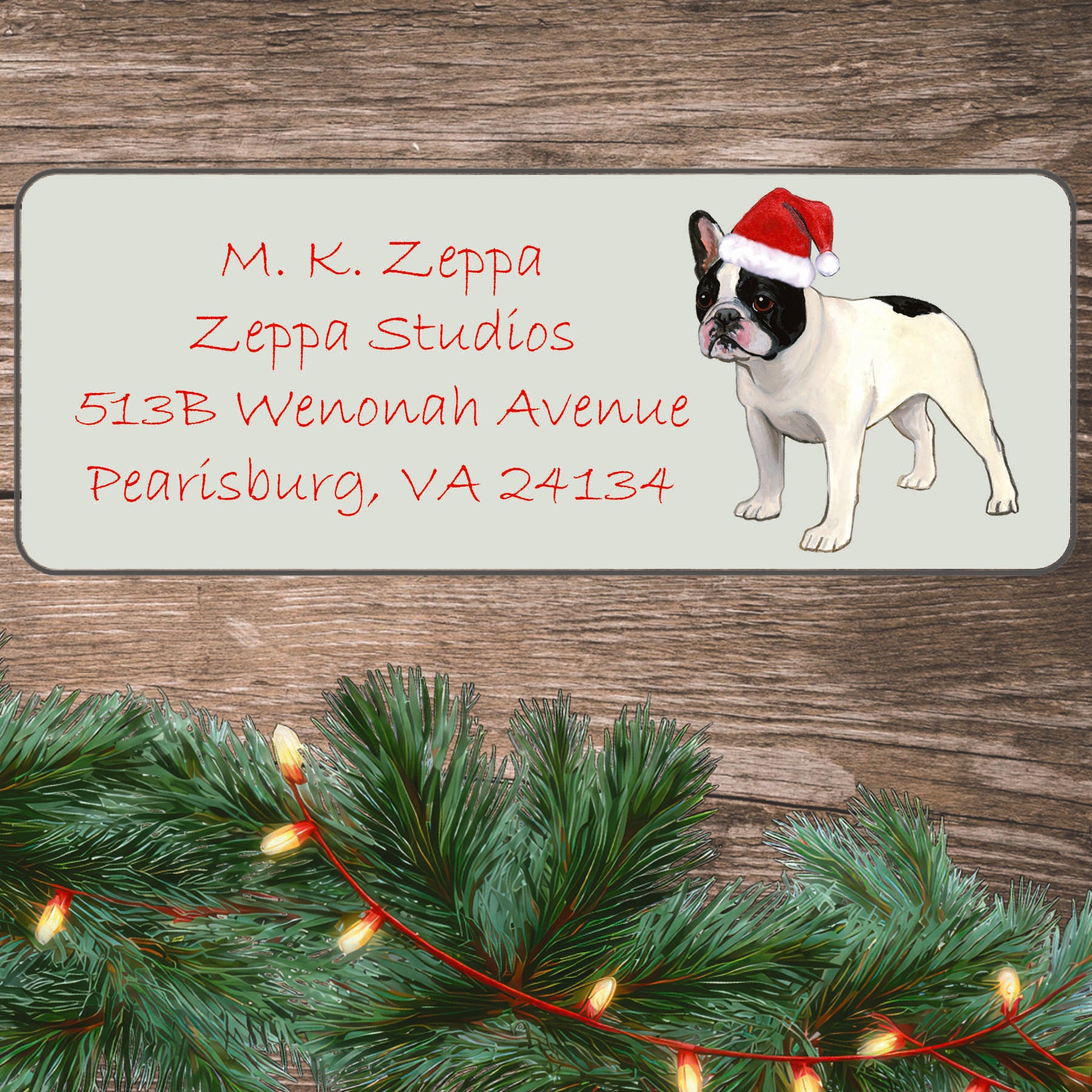 An illustration of a black and white French Bulldog a Santa Hat on an ivory return address label set off by a wood surface with holiday greenery background.