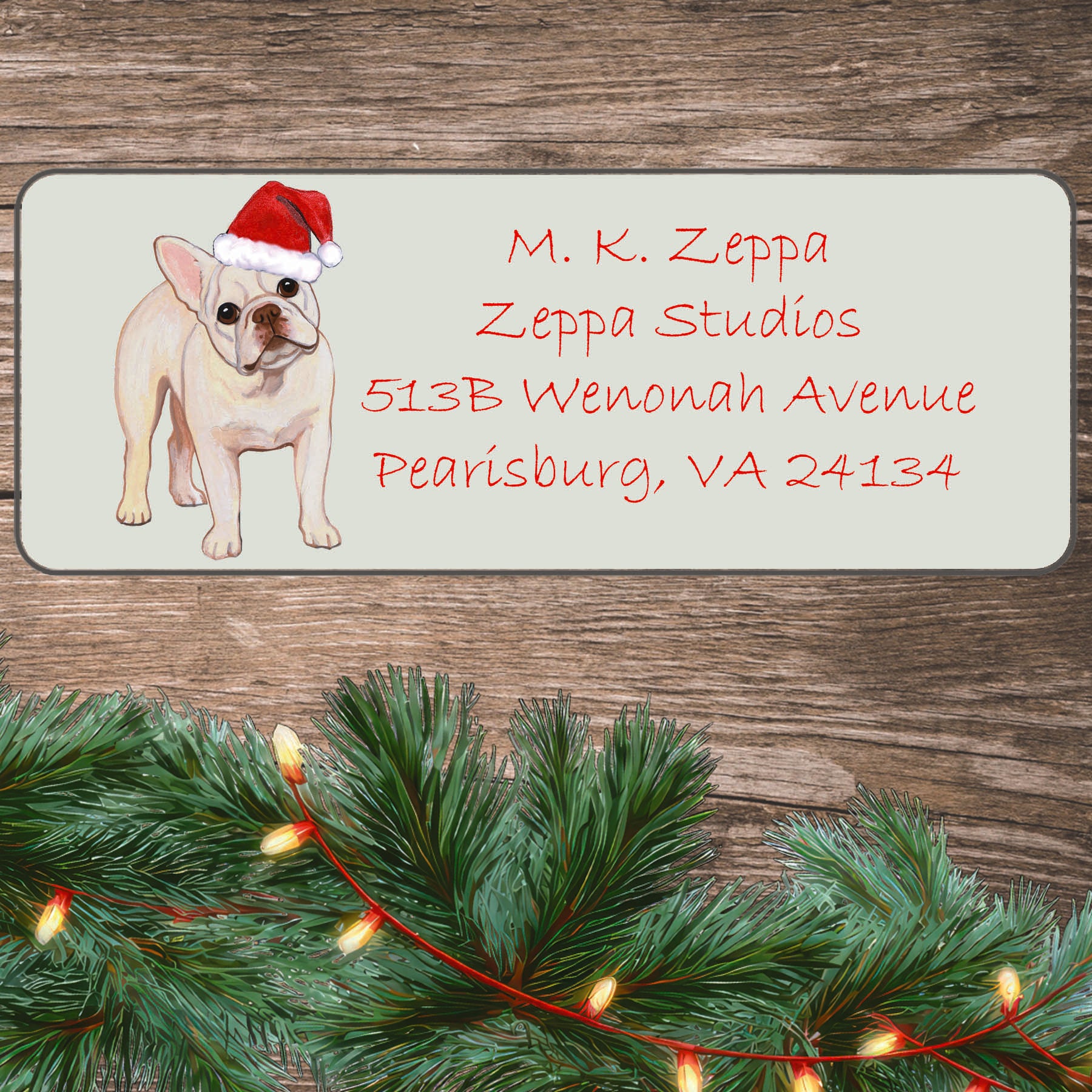 An illustration of a cream French Bulldog  wearing a Santa Hat on an ivory return address label set off by a wood surface with holiday greenery background.