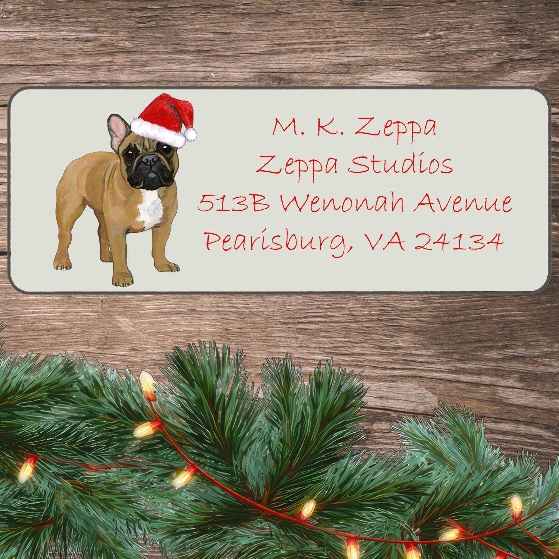 An illustration of a fawn French Bulldog wearing a Santa Hat on an ivory return address label set off by a wood surface with holiday greenery background.