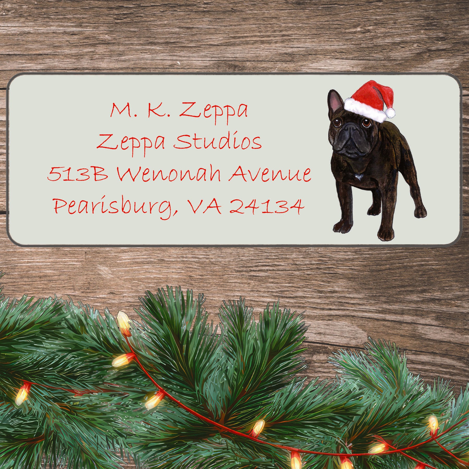 An illustration of a French Bulldog wearing a Santa Hat on an ivory return address label set off by a wood surface with holiday greenery background.