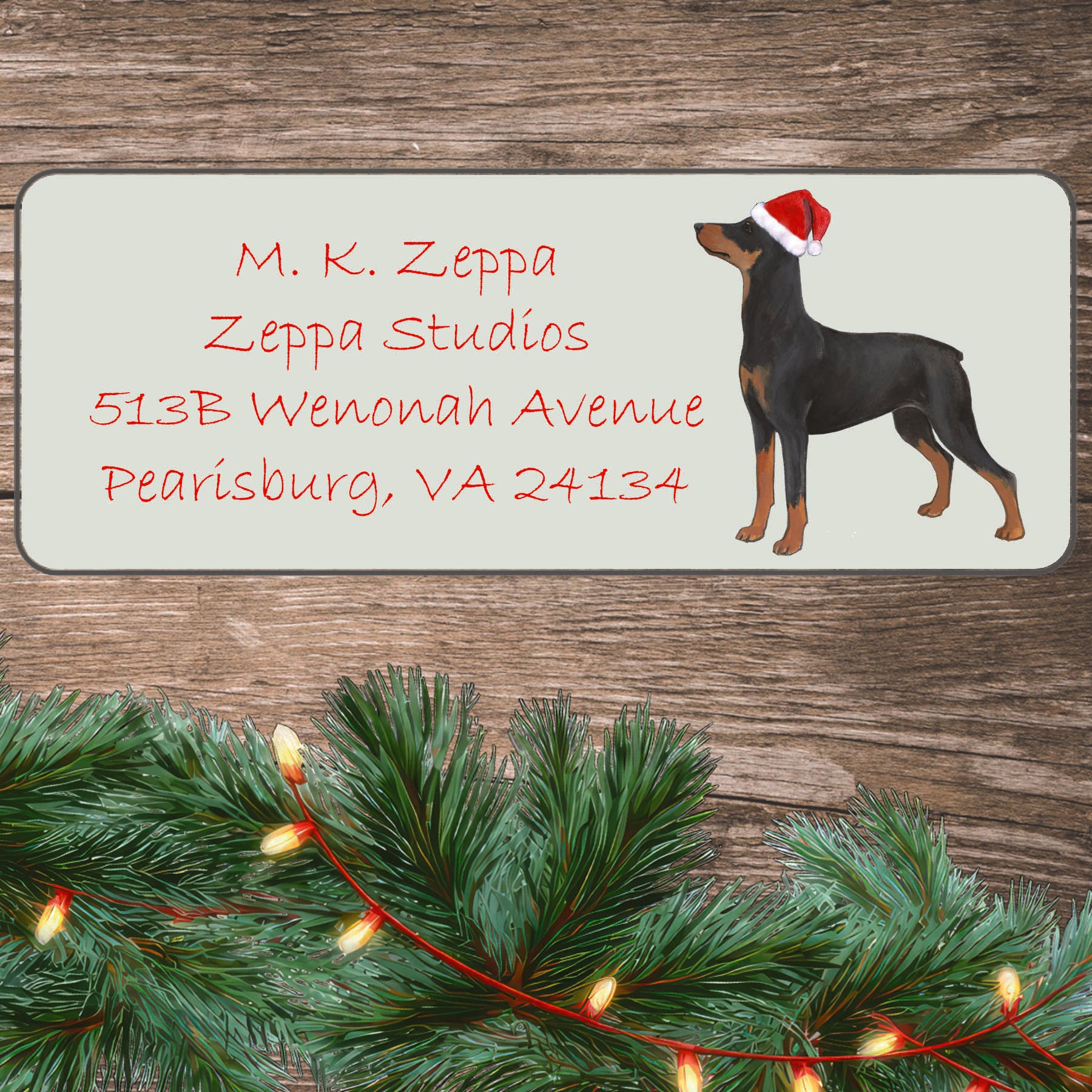 An illustration of a German Pinscher wearing a Santa Hat on an ivory return address label set off by a wood surface with holiday greenery background.