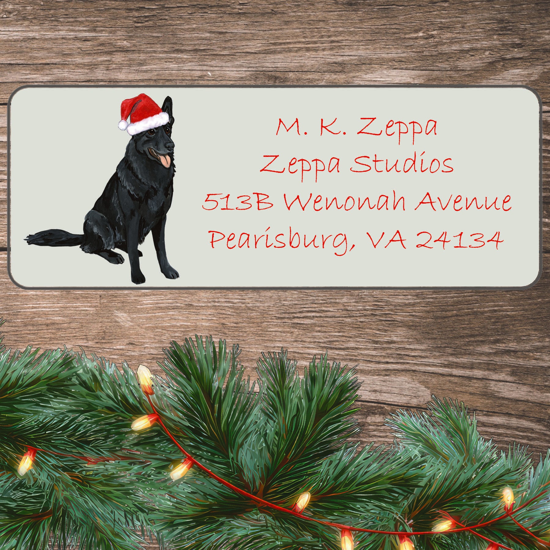 An illustration of a black German Shepherd wearing a Santa Hat on an ivory return address label set off by a wood surface with holiday greenery background.