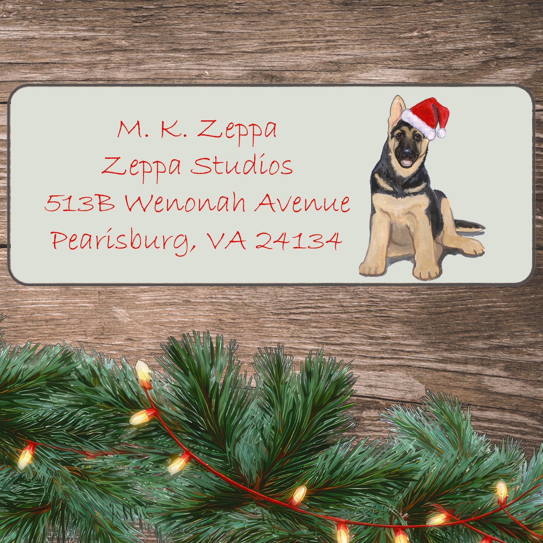 An illustration of a German Shepherd puppy wearing a Santa Hat on an ivory return address label set off by a wood surface with holiday greenery background.