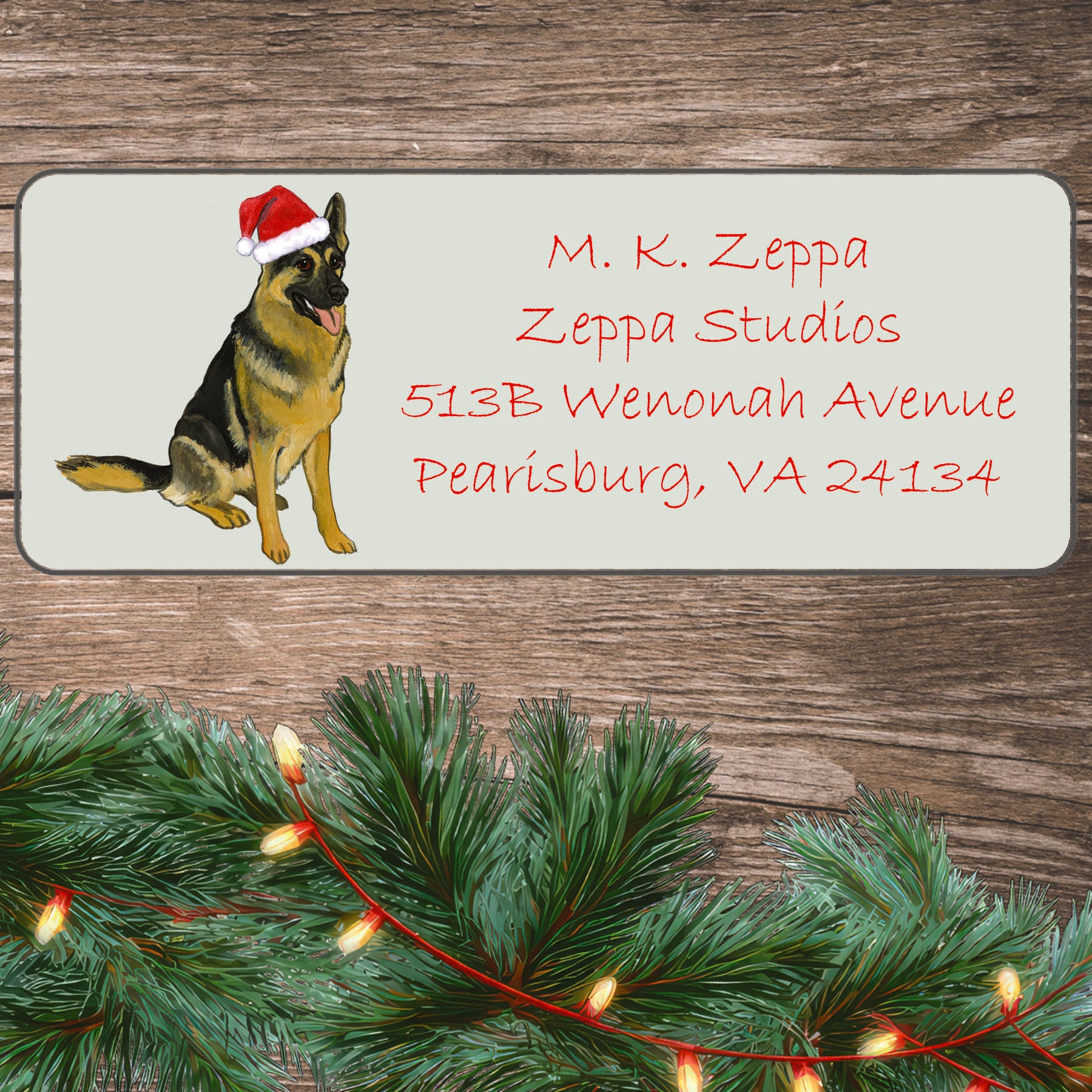 An illustration of a German Shepherd wearing a Santa Hat on an ivory return address label set off by a wood surface with holiday greenery background.