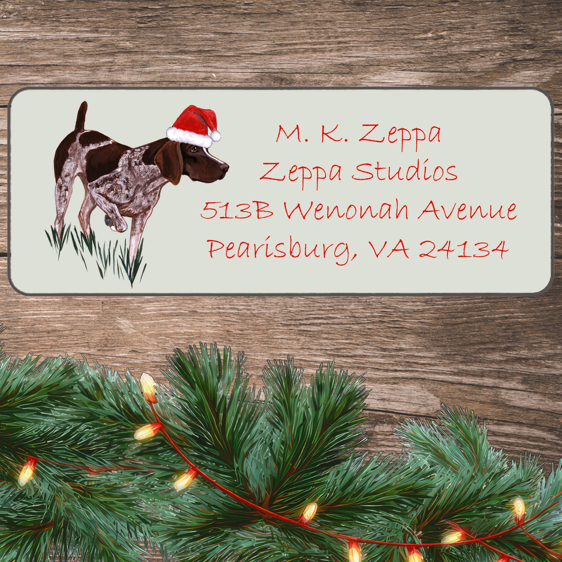 A German Shorthair Pointer in a Santa Hat on an ivory return address label set off by a wood surface with holiday greenery background.