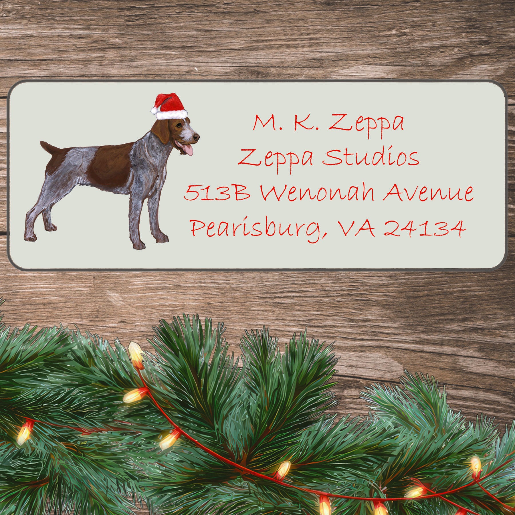 A German WIrehaired Pointer in a Santa Hat on an ivory return address label set off by a wood surface with holiday greenery background.
