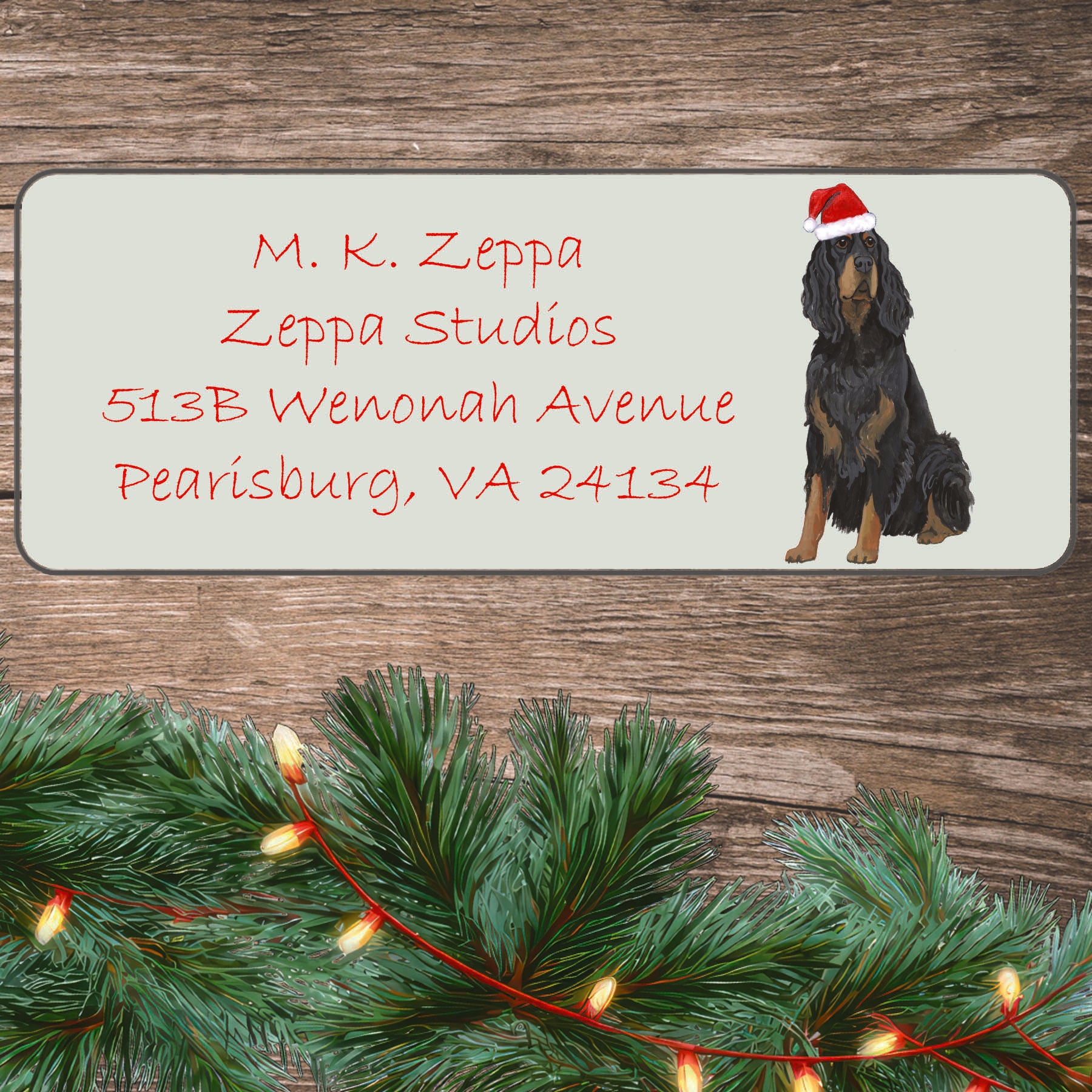 An illustration of a Gordon Setter wearing a Santa Hat on an ivory return address label set off by a wood surface with holiday greenery background.