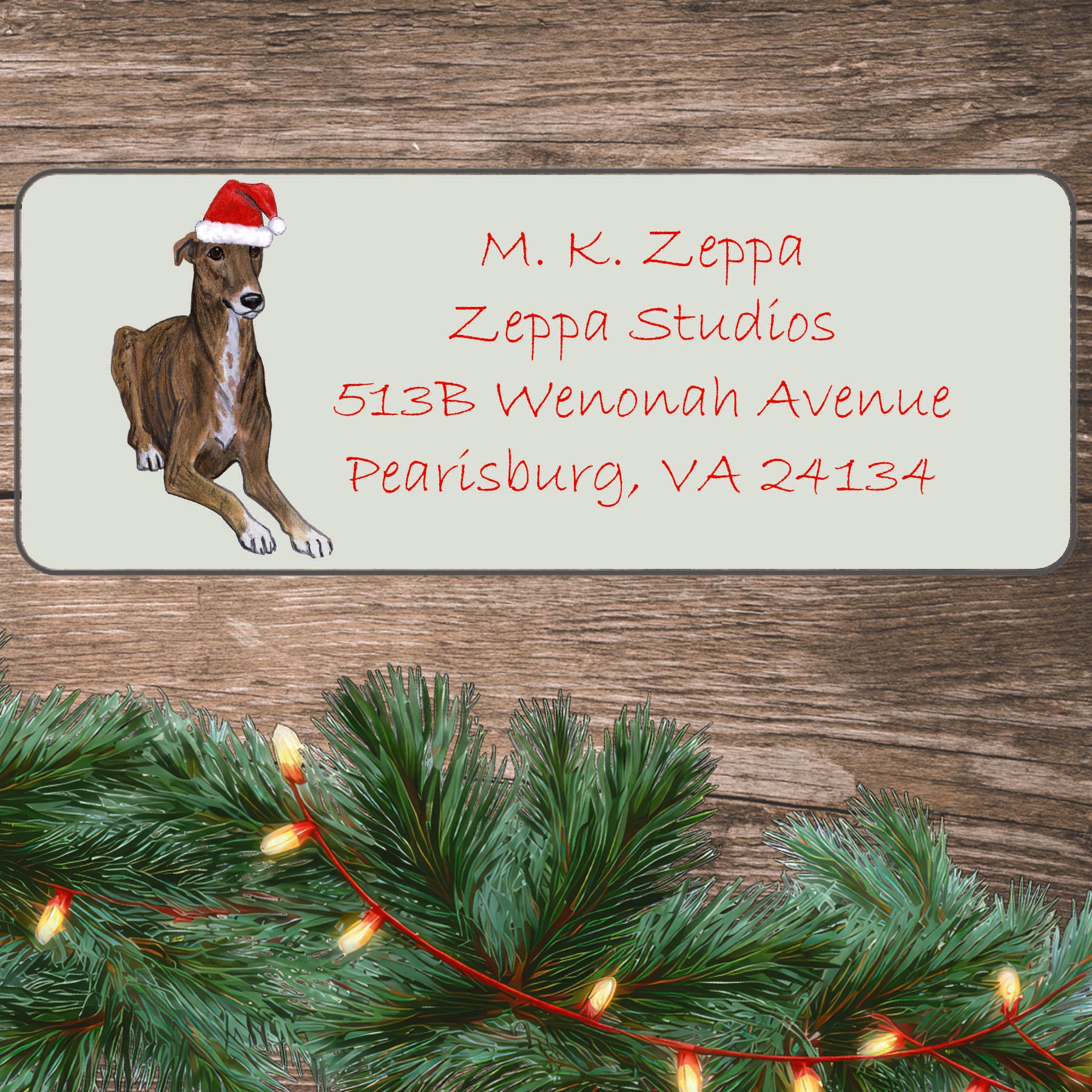 An illustration of a Greyhound wearing a Santa Hat on an ivory return address label set off by a wood surface with holiday greenery background.
