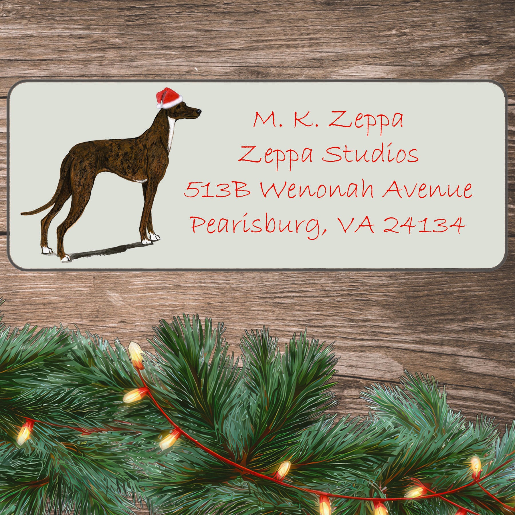 An illustration of a standing Greyhound wearing a Santa Hat on an ivory return address label set off by a wood surface with holiday greenery background.