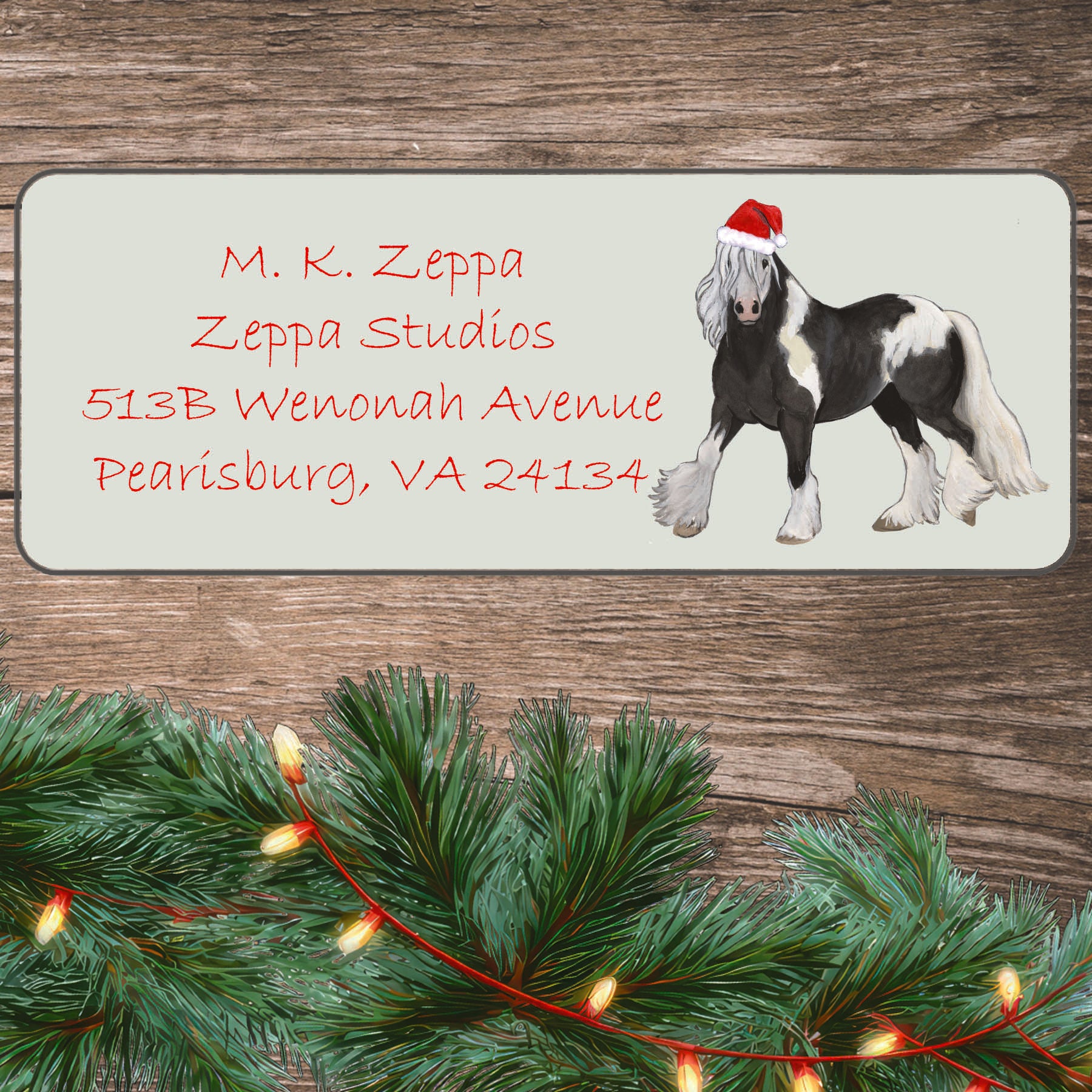A Gypsy Vanner Horse in a Santa Hat illustration on an ivory return address label set off by a wood surface with holiday greenery background.