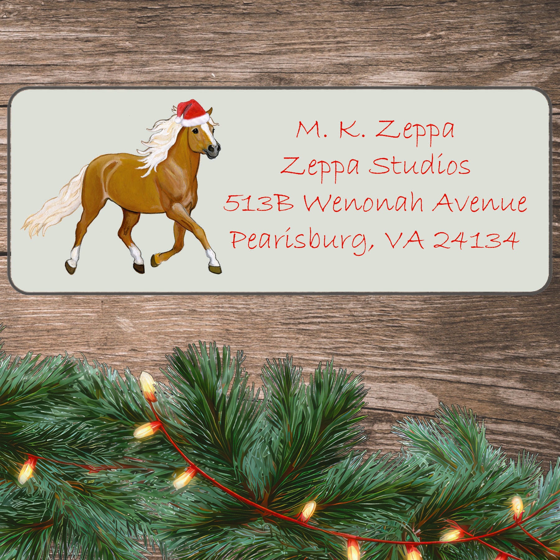 An illustration of a Haflinger Horse wearing a Santa Hat on an ivory return address label set off by a wood surface with holiday greenery background.