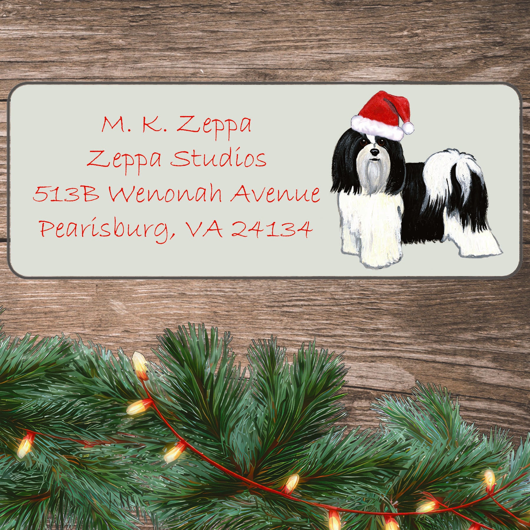 An illustration of a black and white Havanese wearing a Santa Hat on an ivory return address label set off by a wood surface with holiday greenery background.