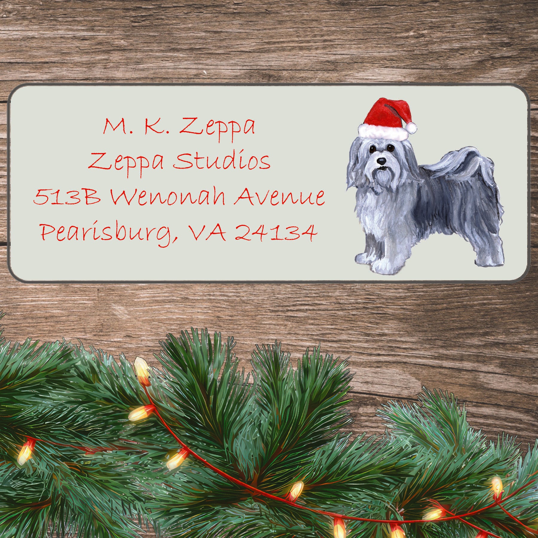 An illustration of a Havanese wearing a Santa Hat on an ivory return address label set off by a wood surface with holiday greenery background.