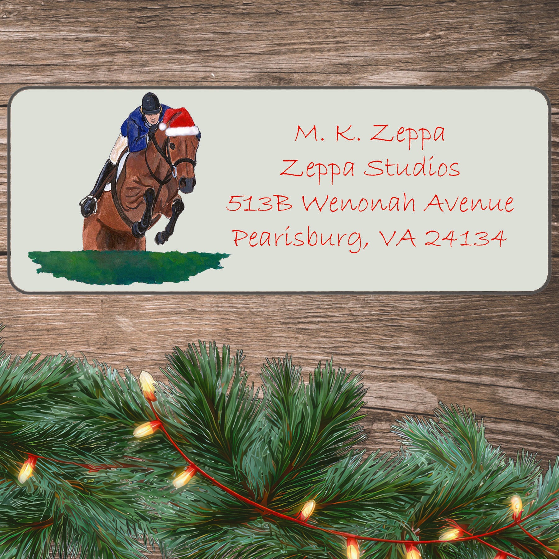 An illustration of a rider on a jumping horse wearing a Santa Hat on an ivory return address label set off by a wood surface with holiday greenery background.