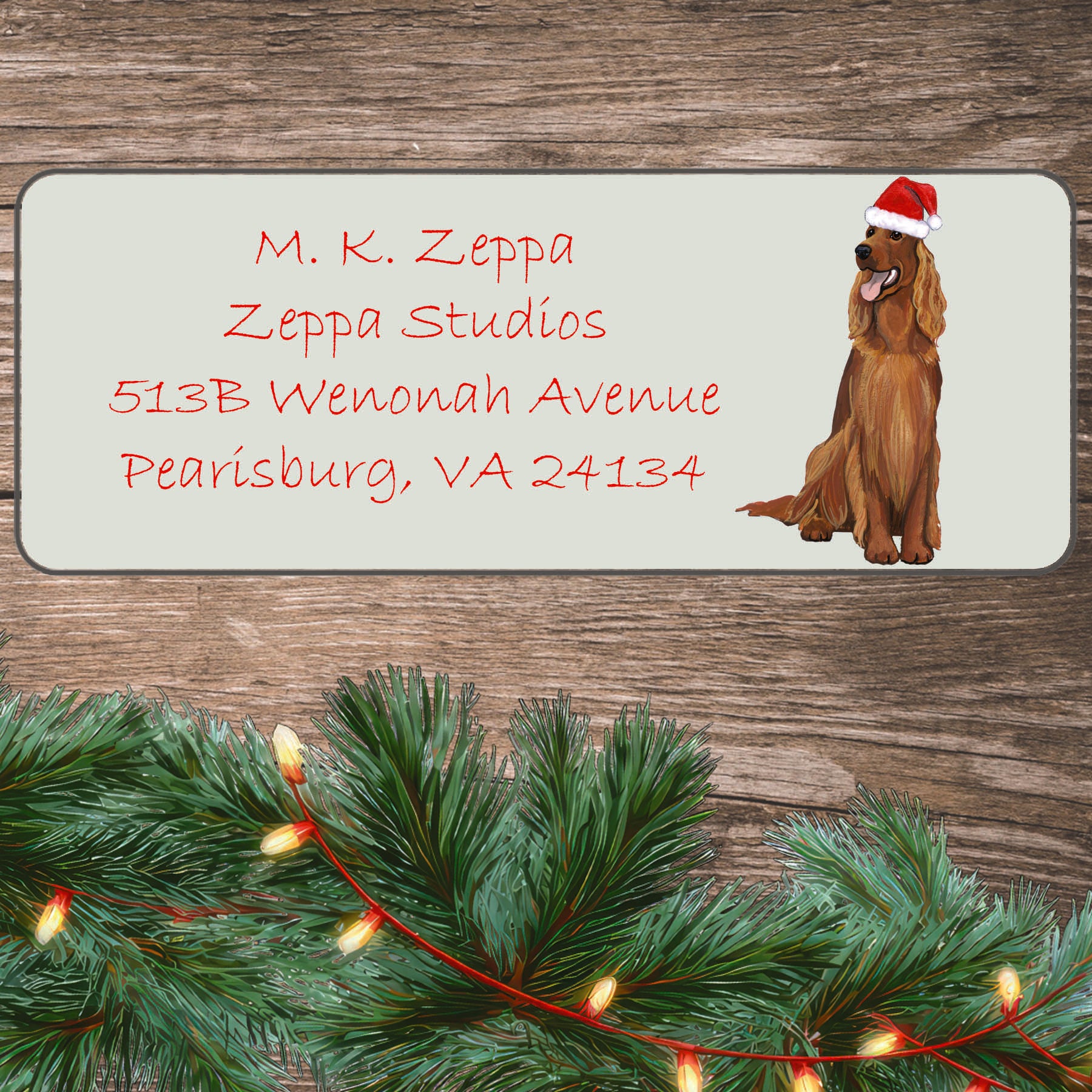 An illustration of an Irish Setter  wearing a Santa Hat on an ivory return address label set off by a wood surface with holiday greenery background.