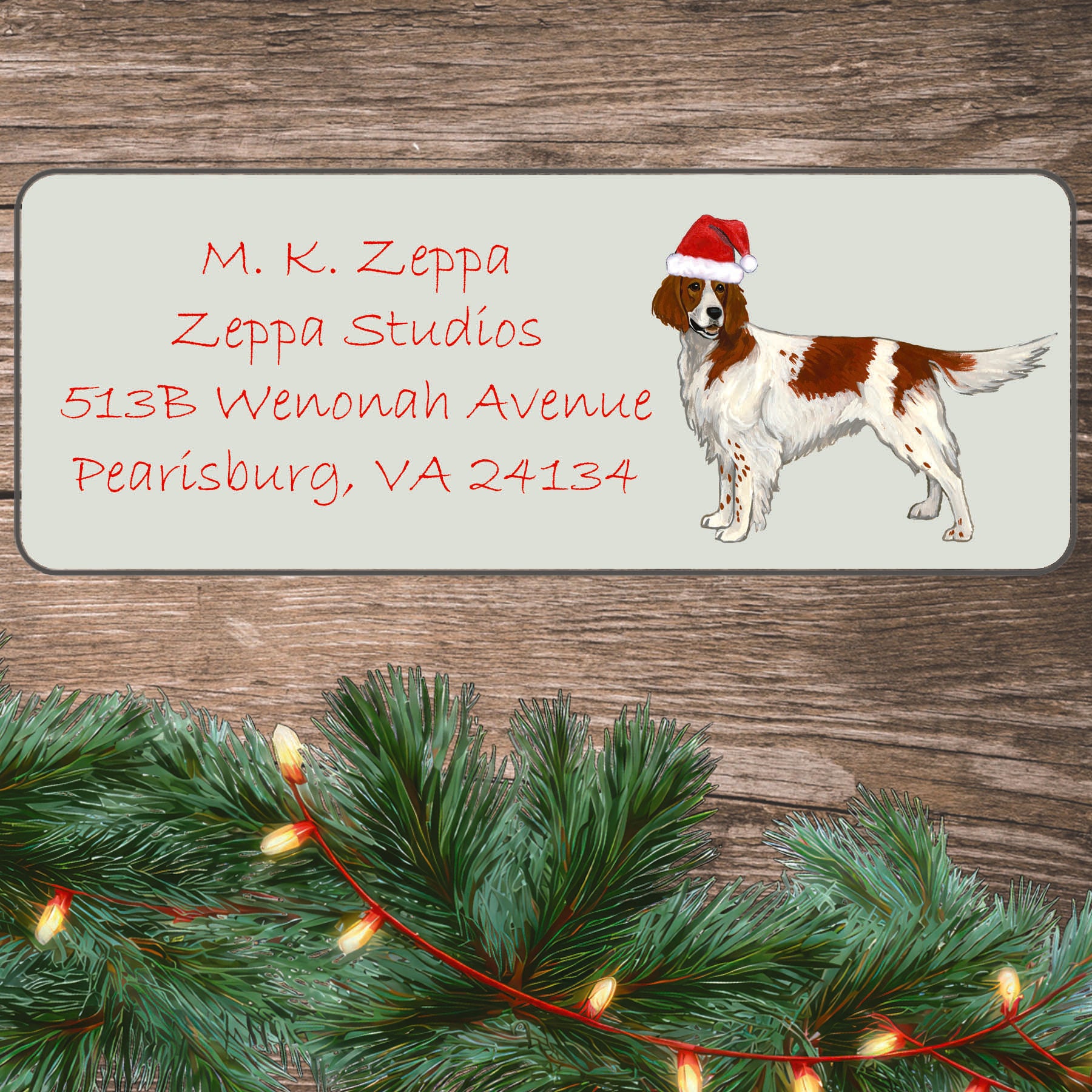 An illustration of an Irish Red and White Setter  wearing a Santa Hat on an ivory return address label set off by a wood surface with holiday greenery background.
