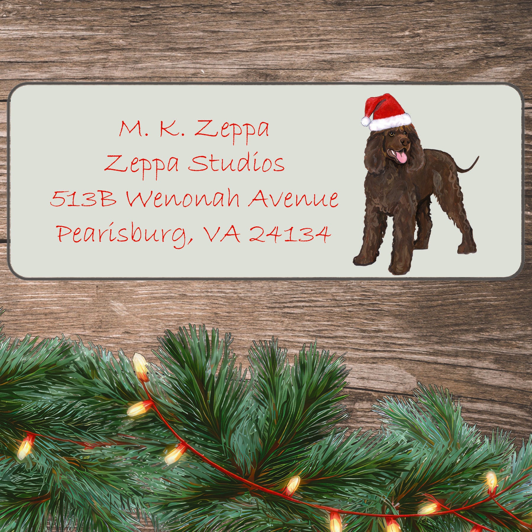 An illustration of an Irish Water Spaniel wearing a Santa Hat on an ivory return address label set off by a wood surface with holiday greenery background.