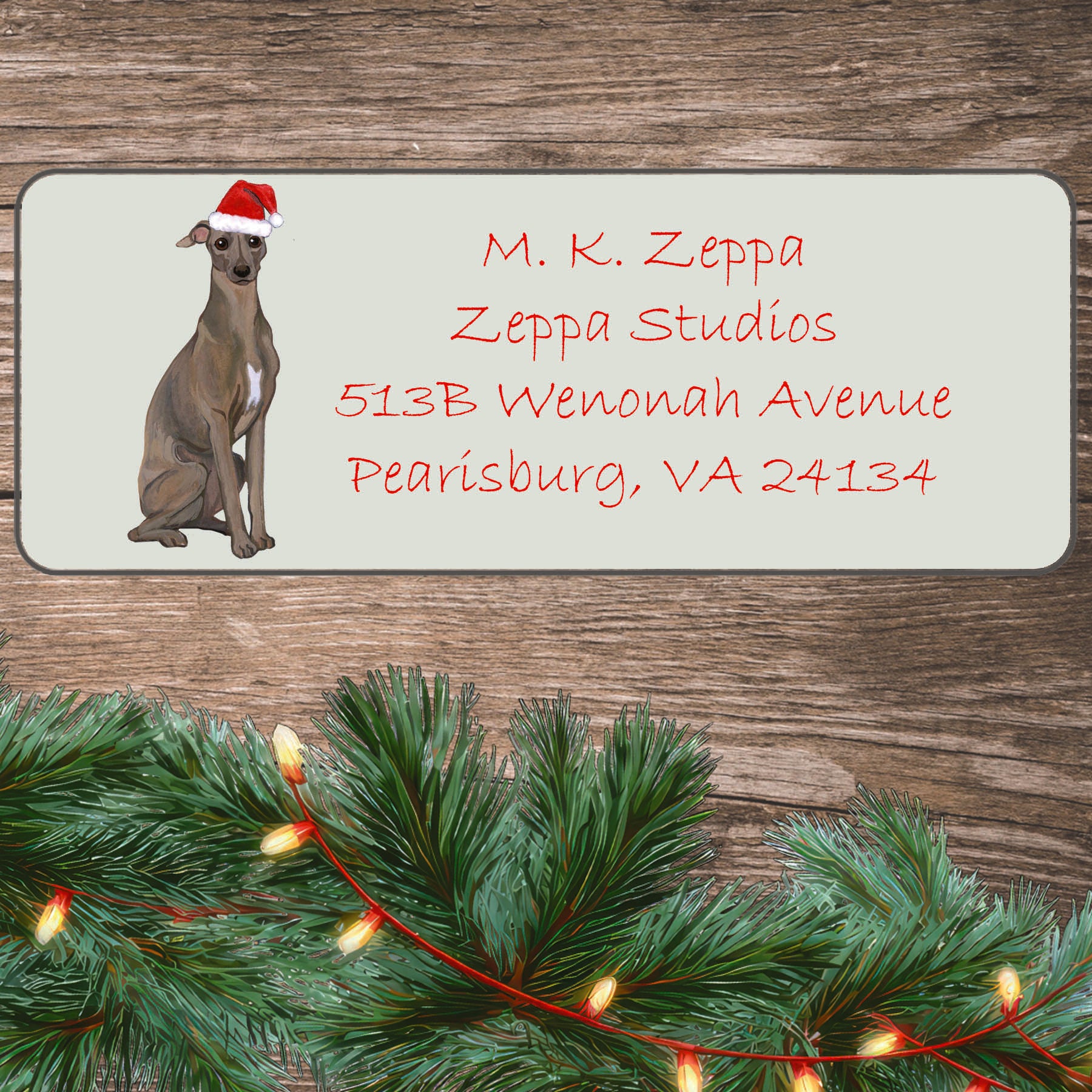 An illustration of a Italian Greyhound wearing a Santa Hat on an ivory return address label set off by a wood surface with holiday greenery background.