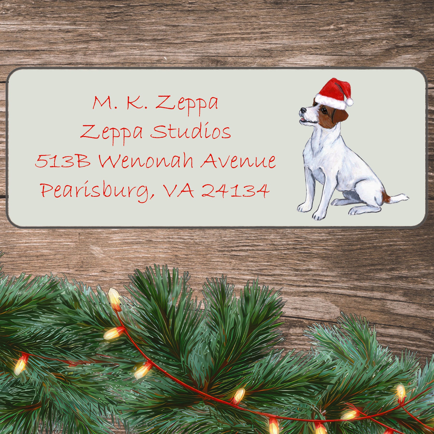 An illustration of a Jack Russell Terrier  wearing a Santa Hat on an ivory return address label set off by a wood surface with holiday greenery background.