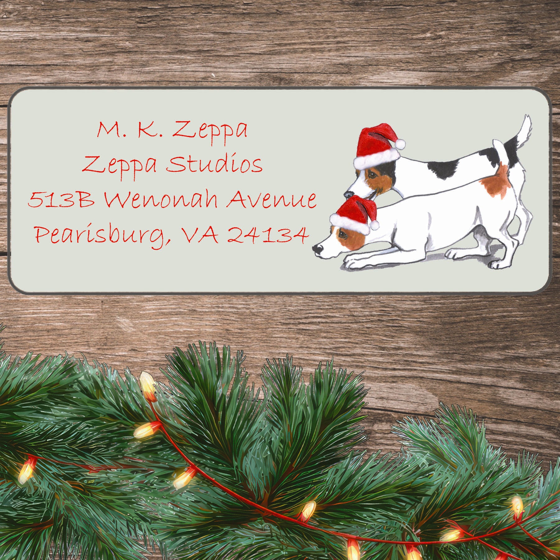 An illustration of a Jack Russell pair wearing a Santa Hat on an ivory return address label set off by a wood surface with holiday greenery background.