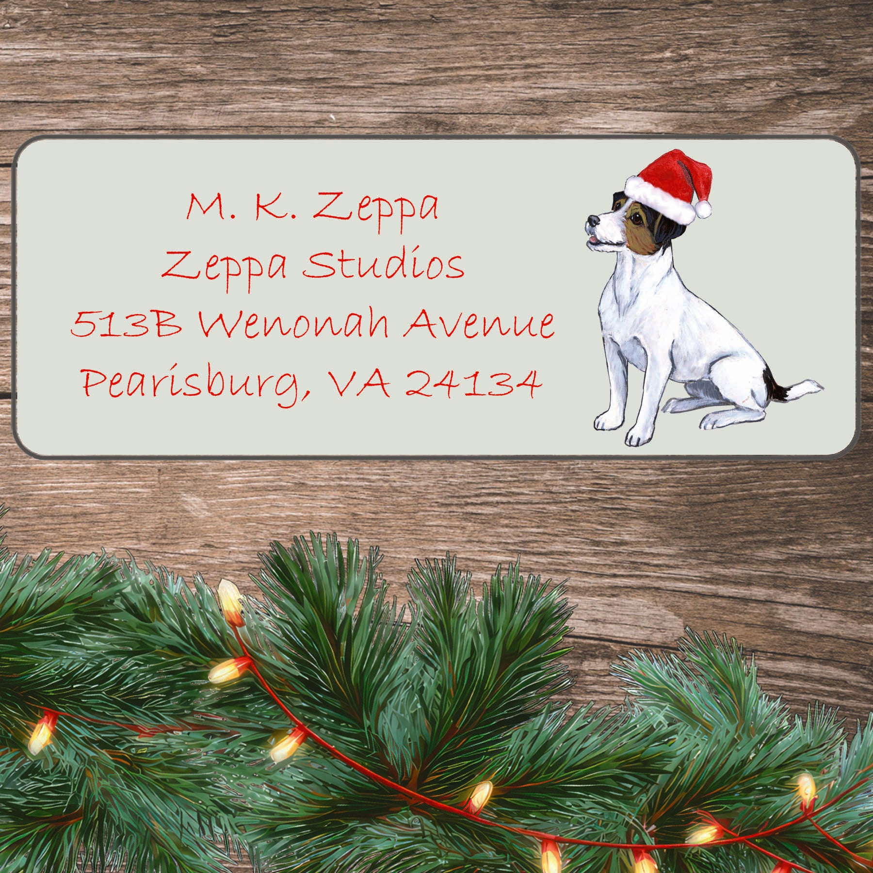 An illustration of a tri color Jack Russell Terrier wearing a Santa Hat on an ivory return address label set off by a wood surface with holiday greenery background.