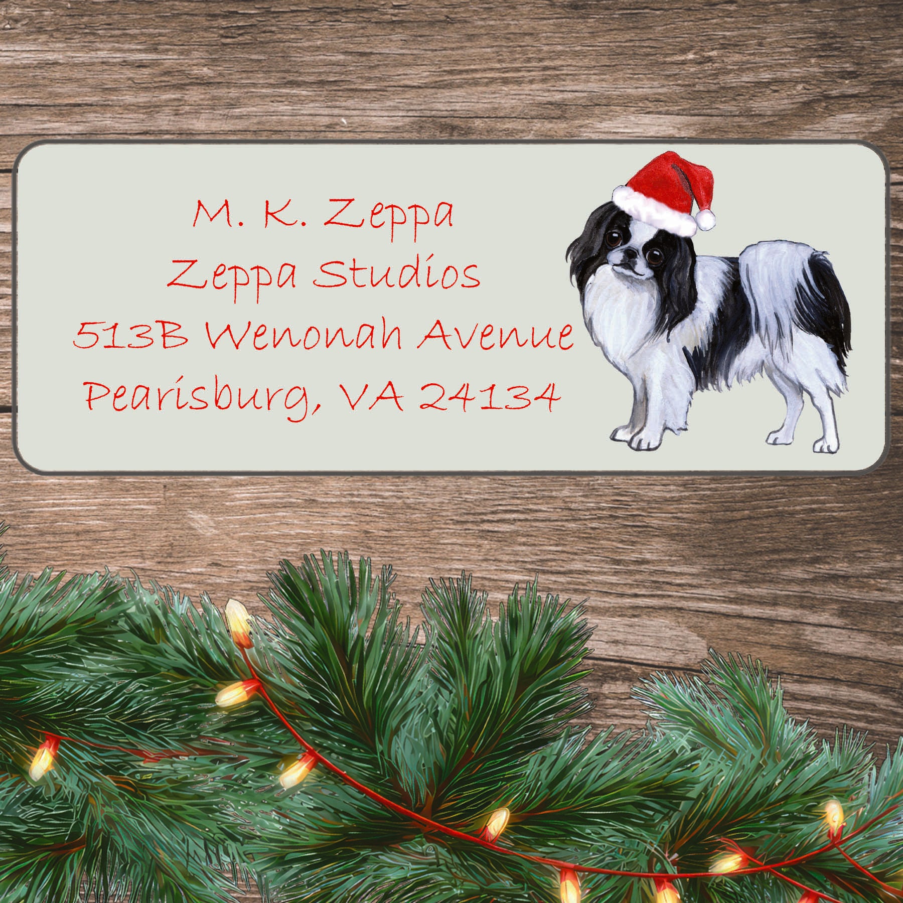 An illustration of a Japanese Chin wearing a Santa Hat on an ivory return address label set off by a wood surface with holiday greenery background.