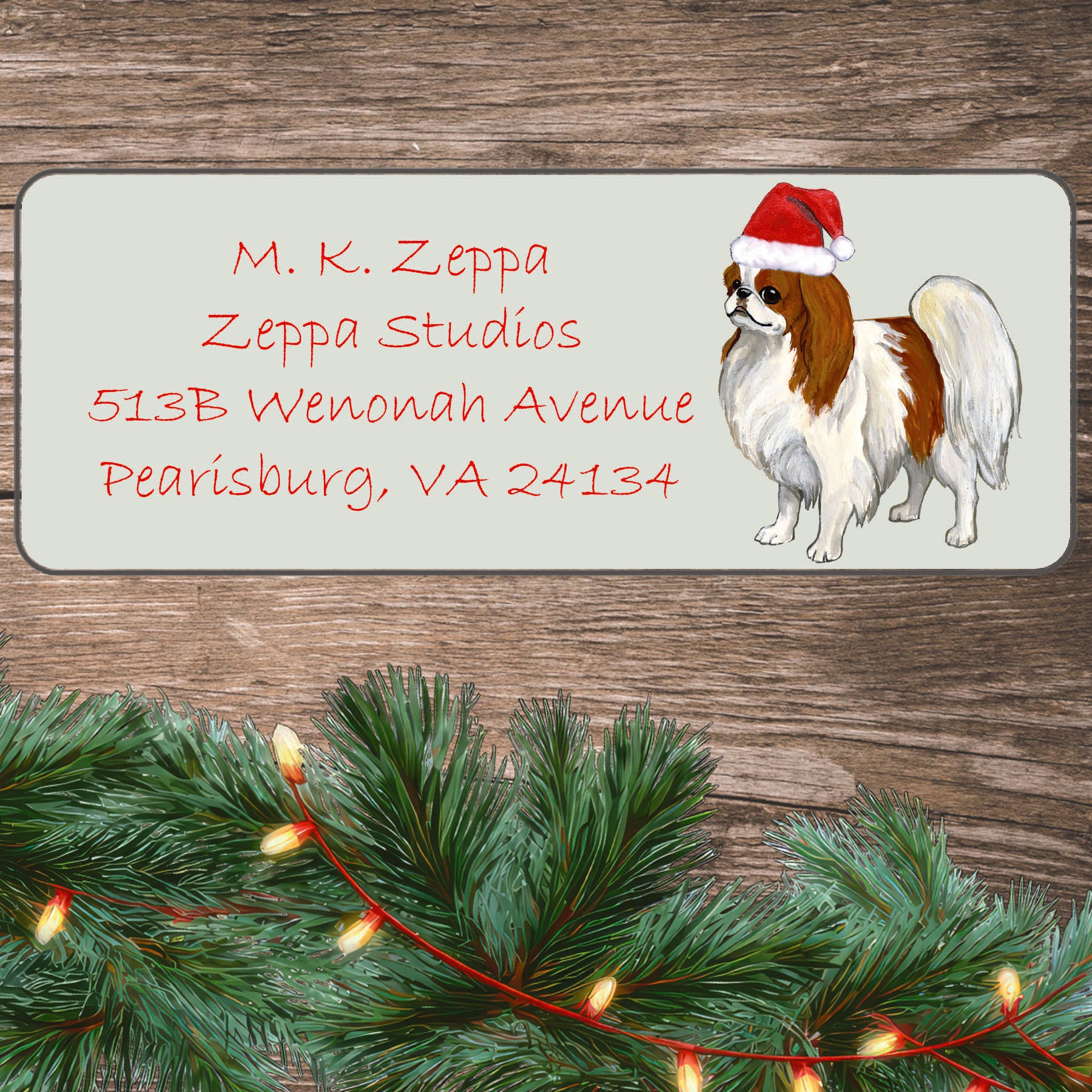 An illustration of a red and white Japanese Chin wearing a Santa Hat on an ivory return address label set off by a wood surface with holiday greenery background.