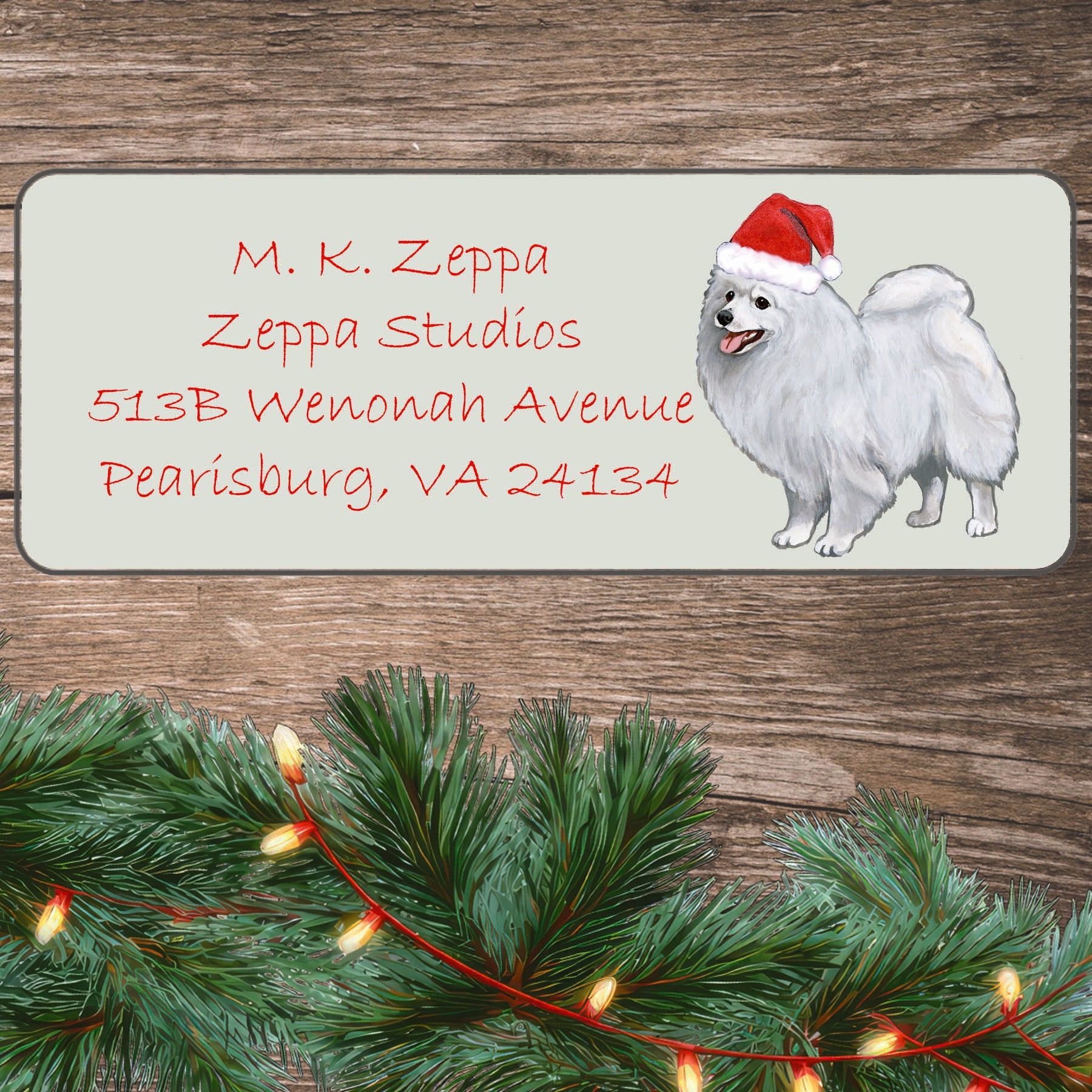 A Japanese Spitz in a Santa Hat illustration on an ivory return address label set off by a wood surface with holiday greenery background.