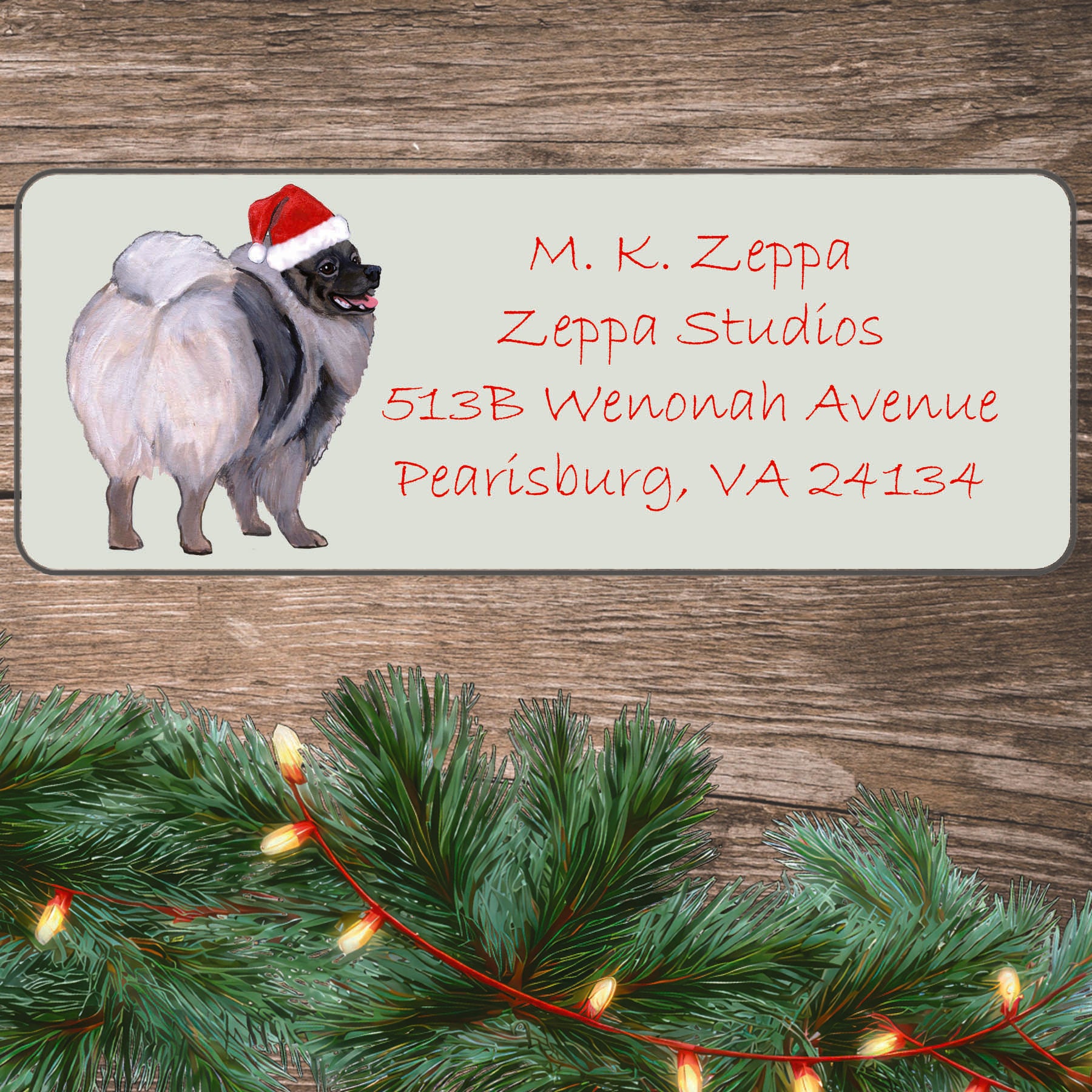 An illustration of a Keeshond wearing a Santa Hat on an ivory return address label set off by a wood surface with holiday greenery background.