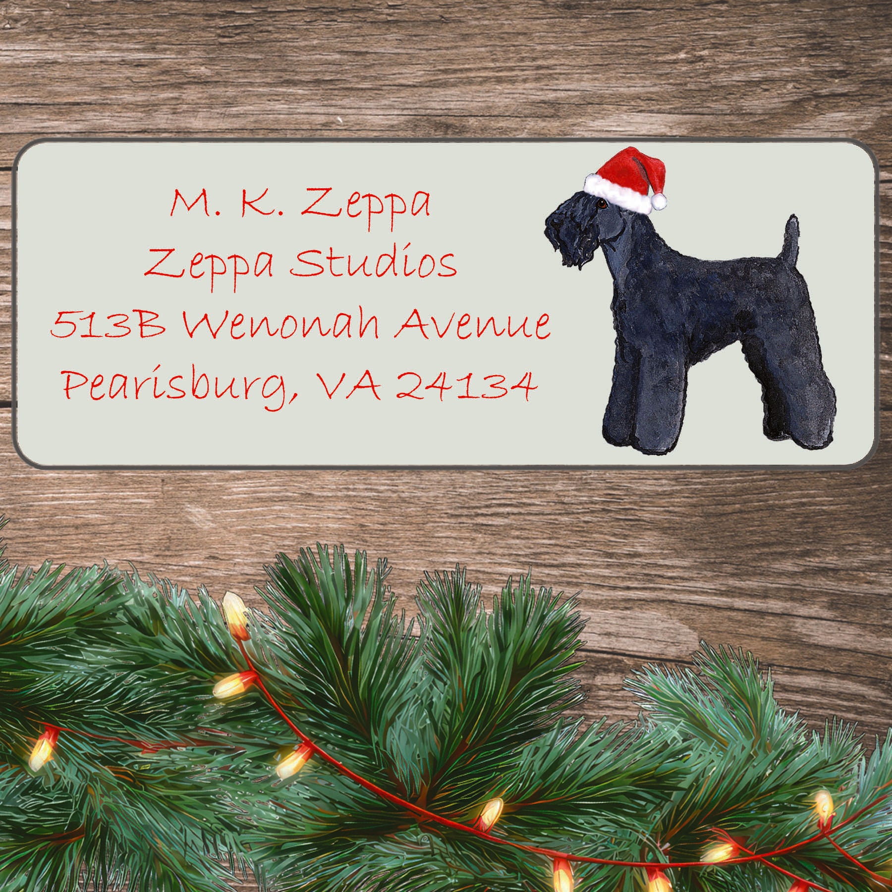 An illustration of a Kerry Blue Terrier wearing a Santa Hat on an ivory return address label set off by a wood surface with holiday greenery background.