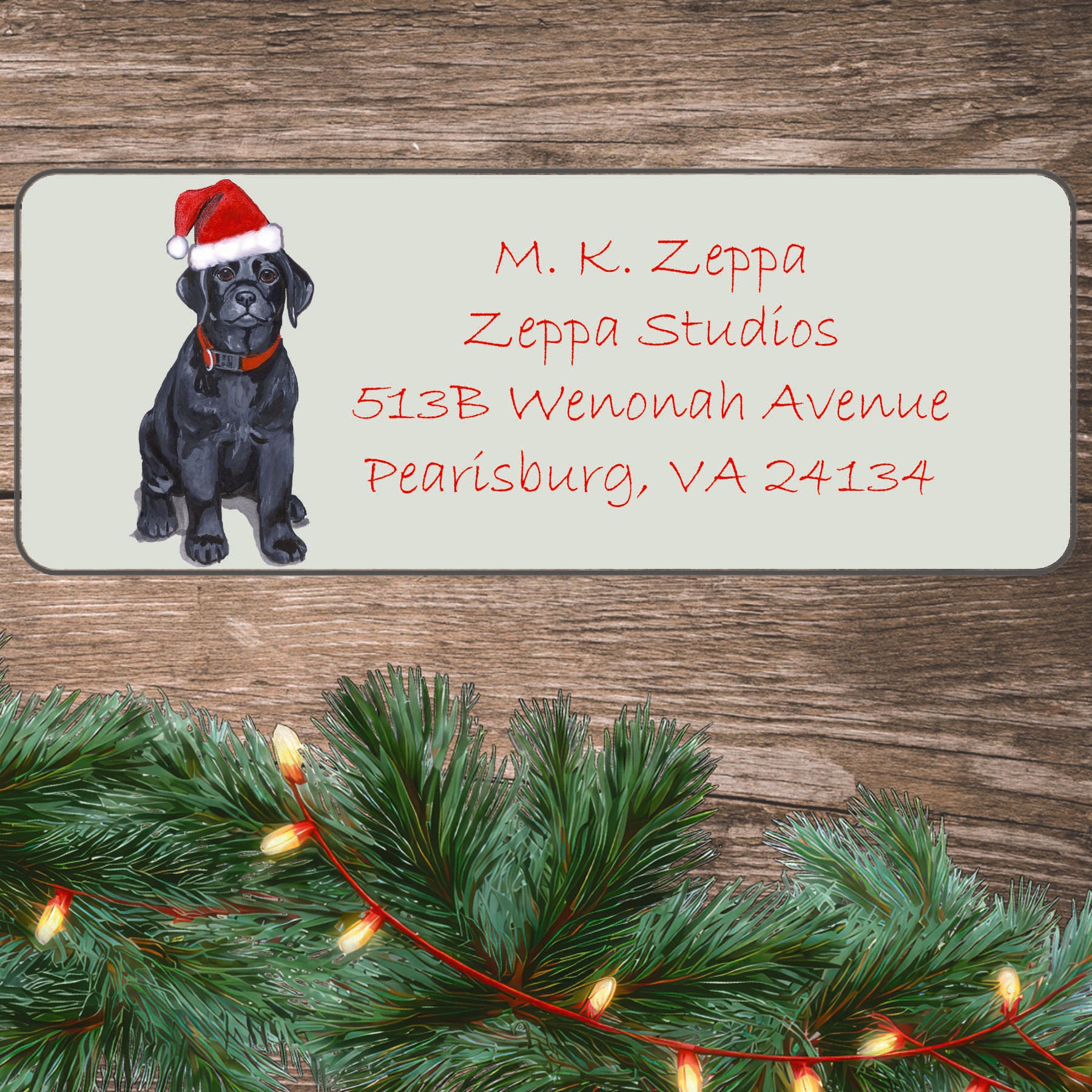 An illustration of a Black Lab puppy wearing a Santa Hat on an ivory return address label set off by a wood surface with holiday greenery background.