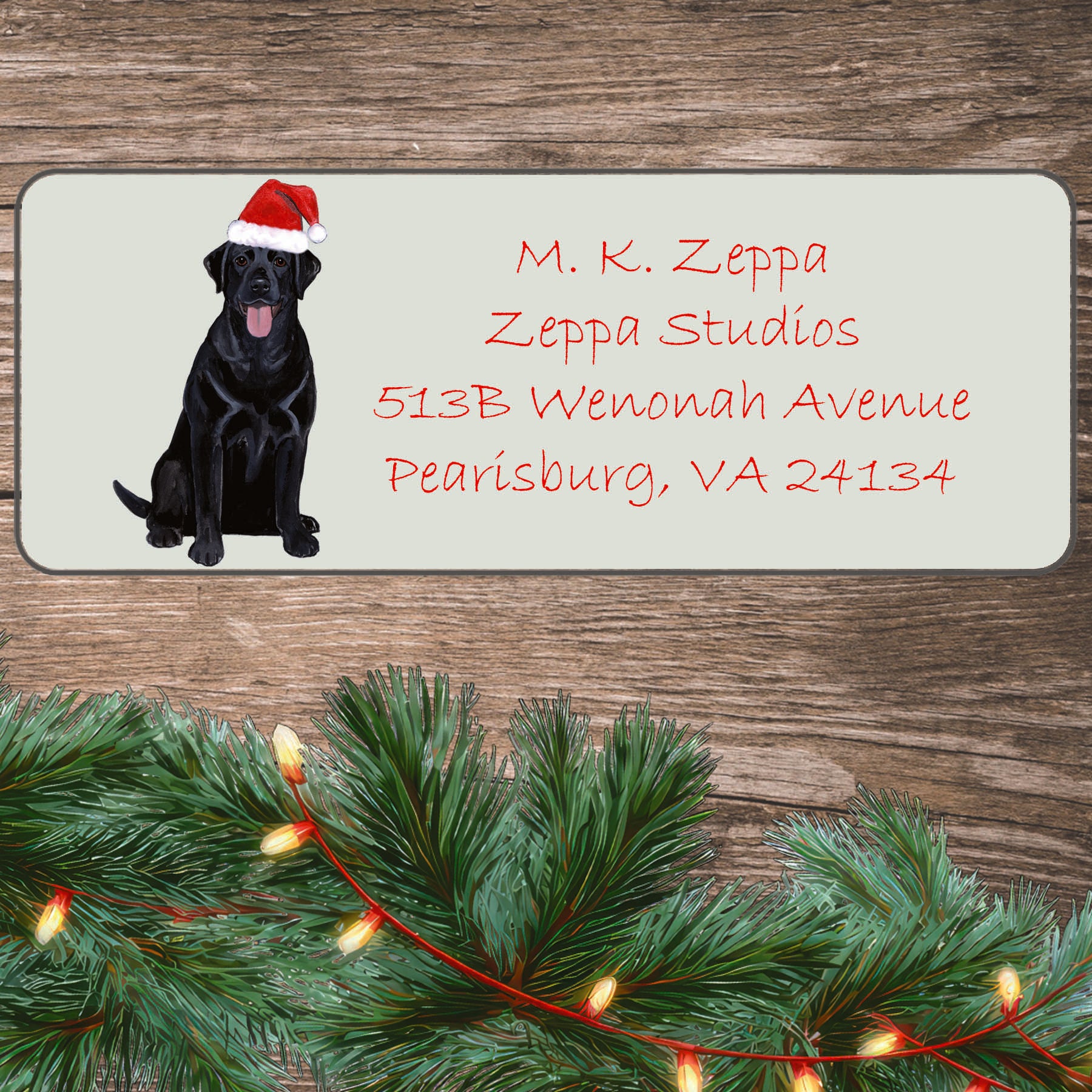 An illustration of a Black Lab wearing a Santa Hat on an ivory return address label set off by a wood surface with holiday greenery background.