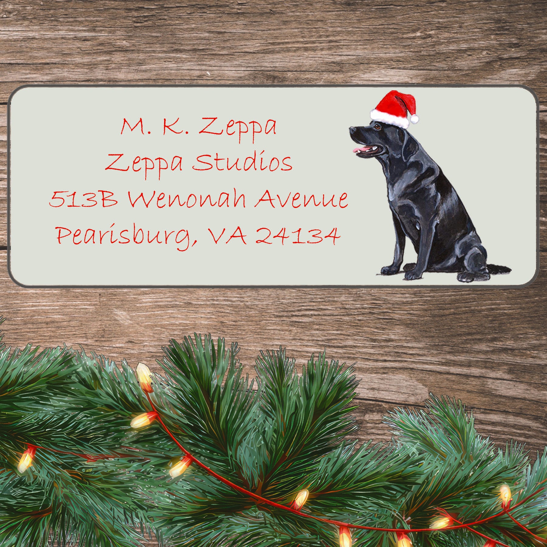 An illustration of a Black Lab sitting wearing a Santa Hat on an ivory return address label set off by a wood surface with holiday greenery background.