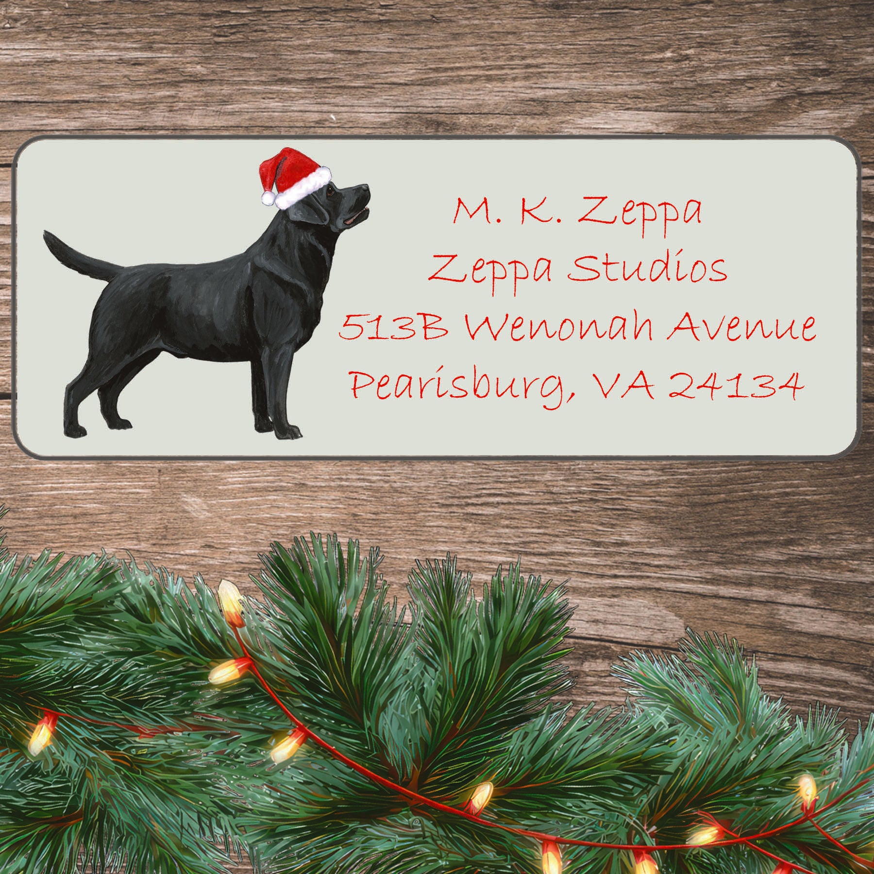 An illustration of a Black Lab standing wearing a Santa Hat on an ivory return address label set off by a wood surface with holiday greenery background.