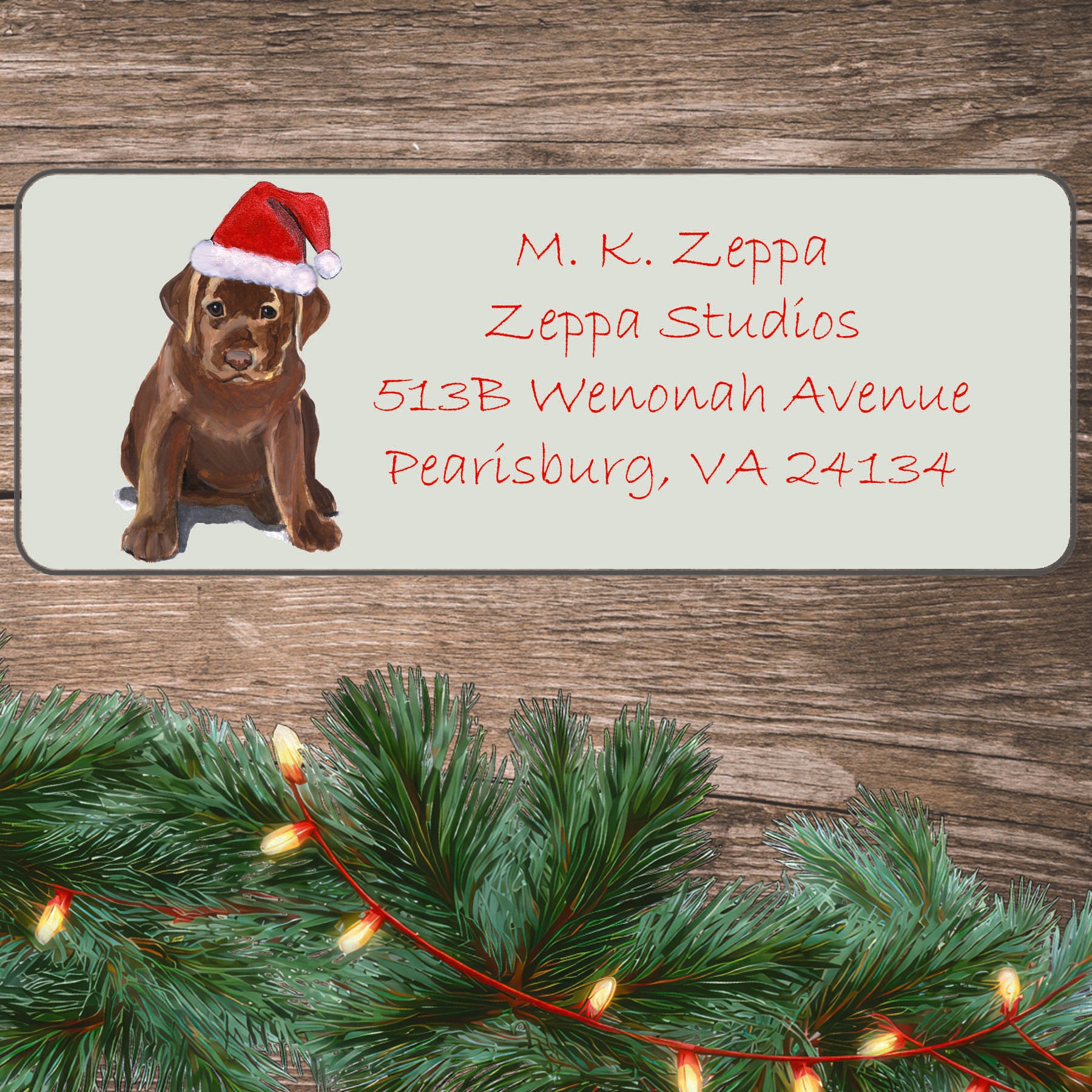 An illustration of a Chocolate Lab Puppy wearing a Santa Hat on an ivory return address label set off by a wood surface with holiday greenery background.