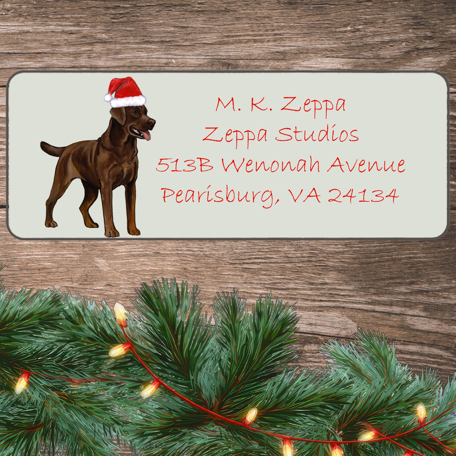 An illustration of a Chocolate Labrador wearing a Santa Hat on an ivory return address label set off by a wood surface with holiday greenery background.