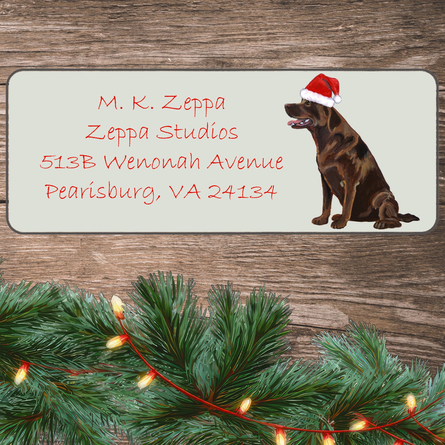 An illustration of a sitting Chocolate Lab wearing a Santa Hat on an ivory return address label set off by a wood surface with holiday greenery background.