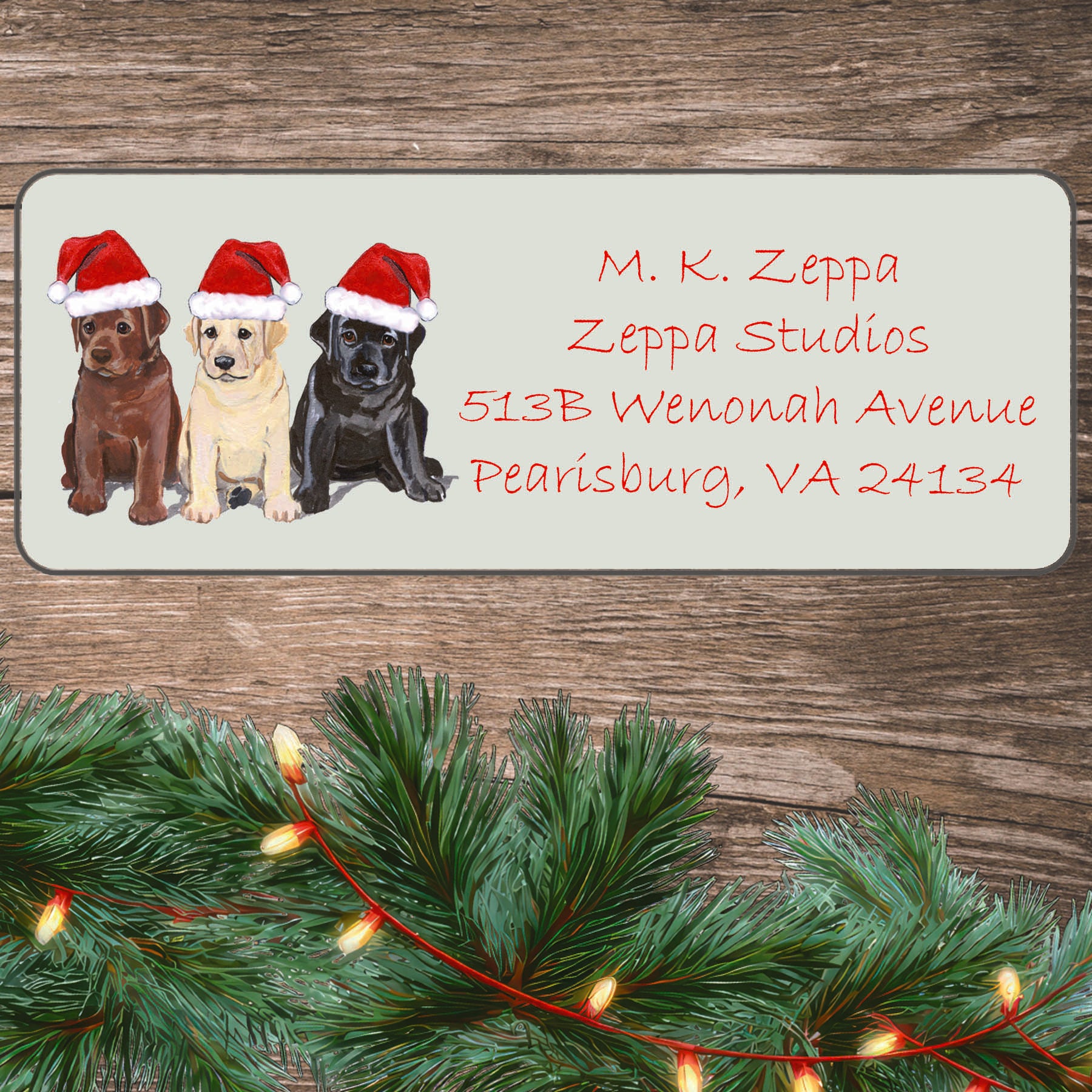 An illustration of 3 Lab Puppies wearing Santa Hats on an ivory return address label set off by a wood surface with holiday greenery background.