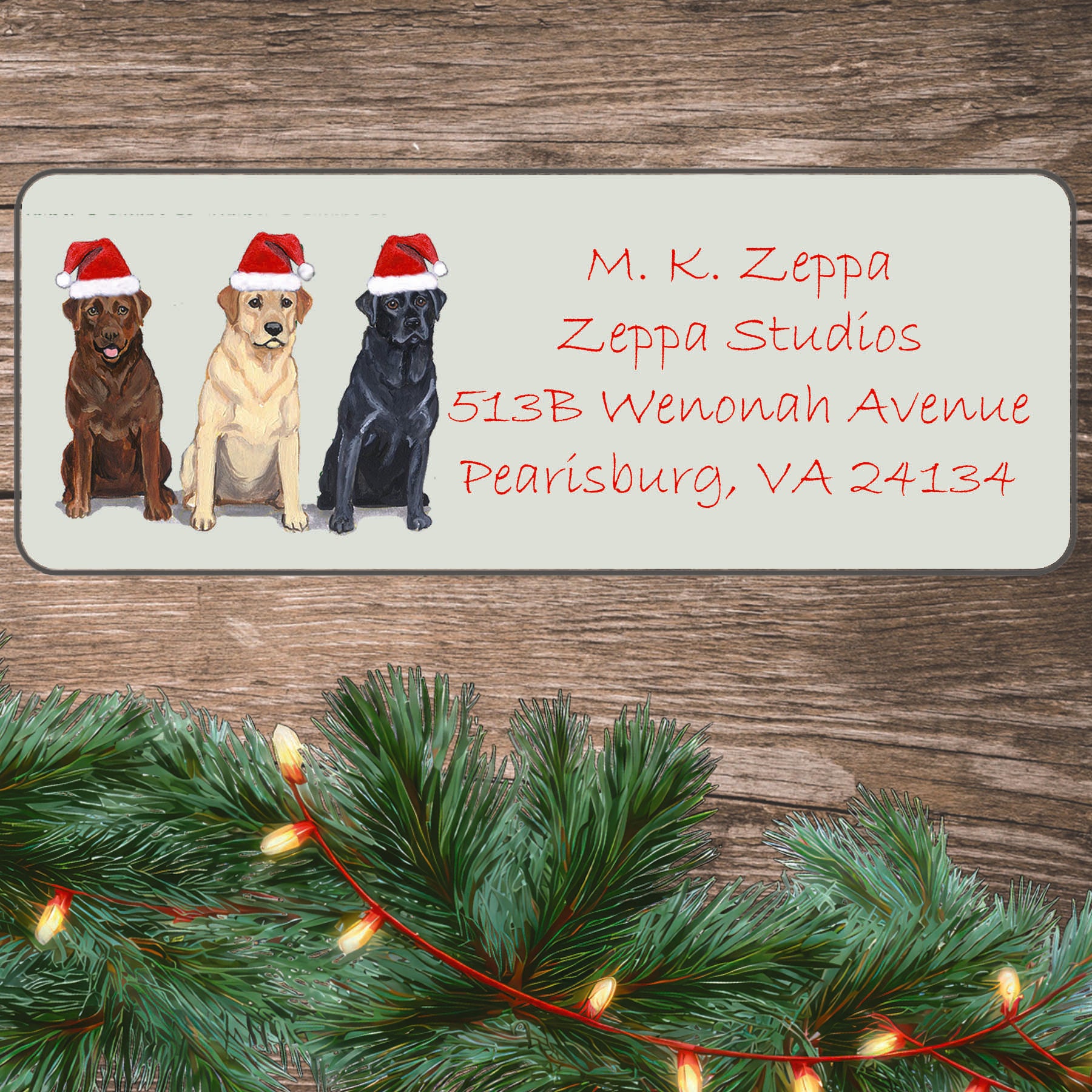 An illustration of 3 Labradors wearing Santa Hats on an ivory return address label set off by a wood surface with holiday greenery background.