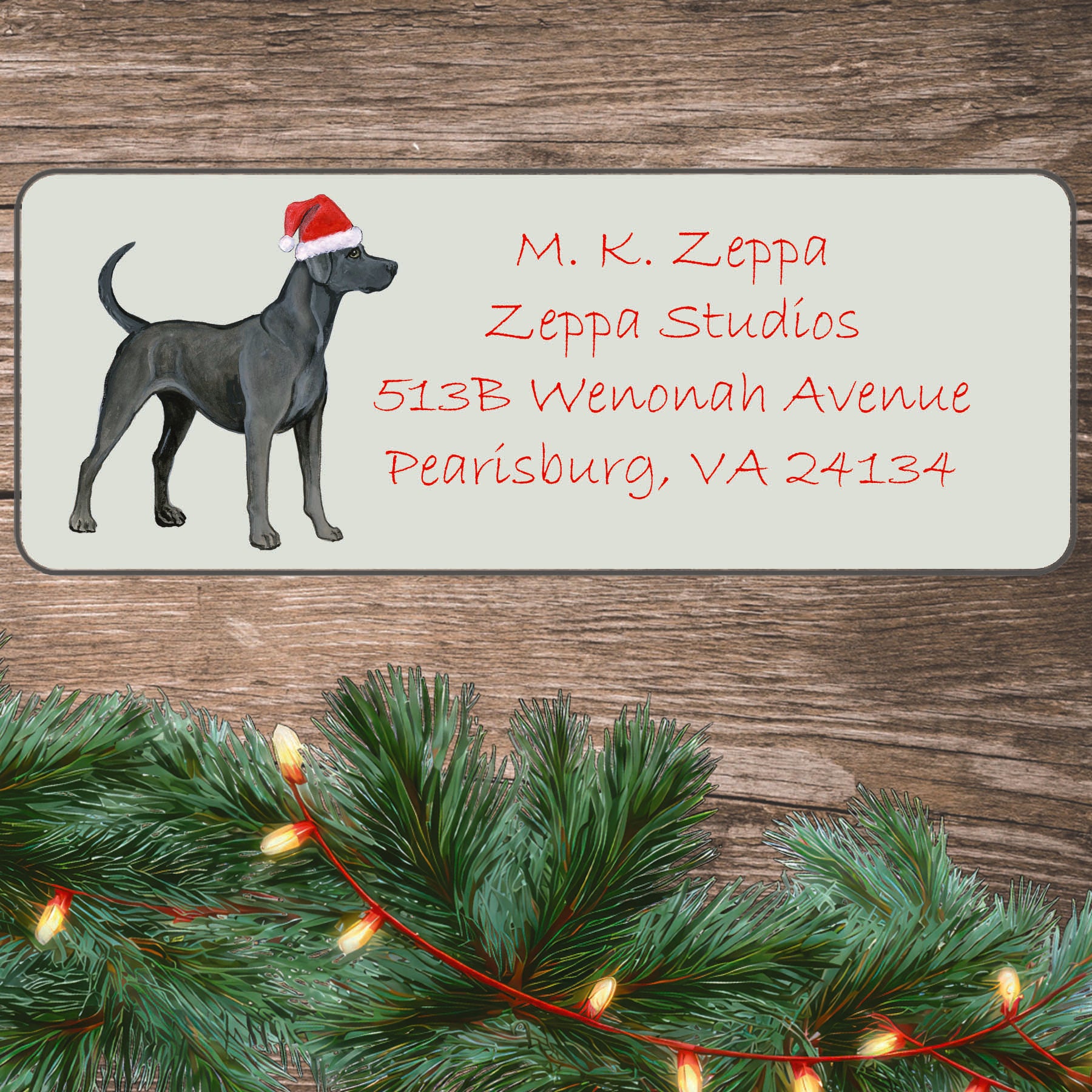 An illustration of a Blue Lacy Dog wearing a Santa Hat on an ivory return address label set off by a wood surface with holiday greenery background.